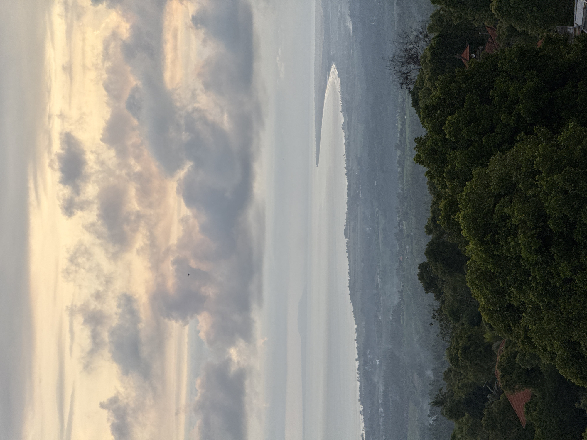 The image shows a landscape with a body of water, possibly a lake or a bay, in the center. On the left side, there is a cloudy sky with varying shades of gray and hints of yellow from the setting sun. The water appears calm and reflects the light from the sky. On the right side, a hillside is covered with lush green trees and vegetation. A few rooftops are visible among the trees, suggesting the presence of buildings. The scene appears to be taken from a high vantage point, offering a panoramic view of the natural landscape and human settlements. The location is Unknown, Unknown.