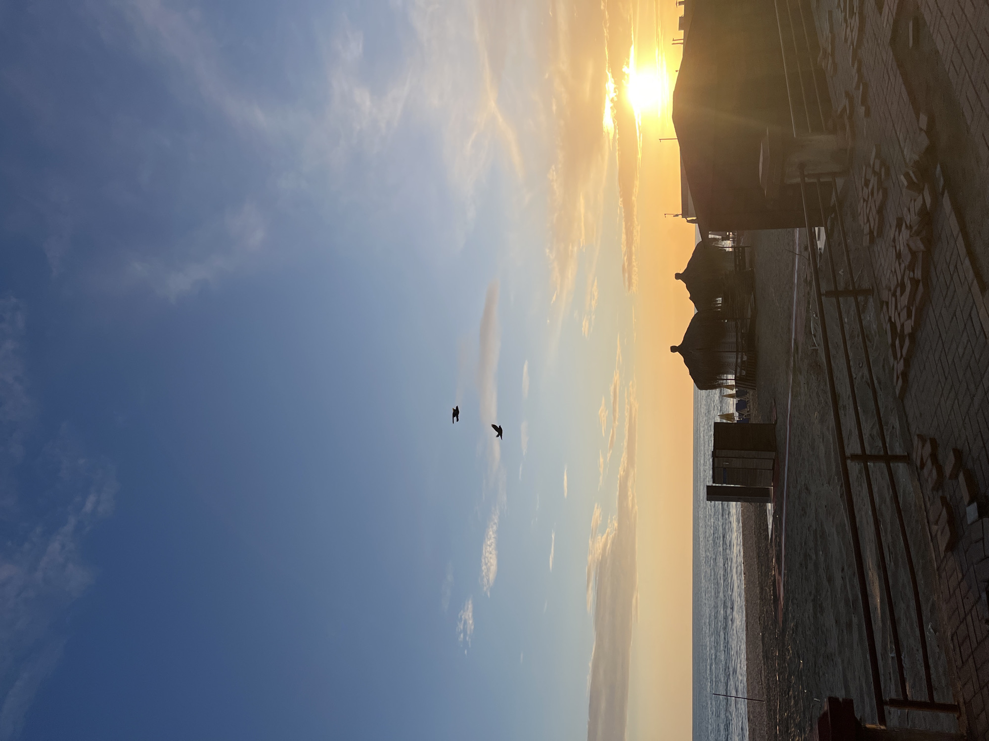The image shows a beach scene at sunset. The sky is a gradient of blue to orange, with the sun partially obscured by clouds near the horizon. Two birds are flying in the sky, silhouetted against the bright background. On the right side of the image, there are beach umbrellas, beach furniture, and a building. The ground in the foreground appears to be a brick surface with some loose bricks. The sea is visible on the right, bordering the beach. This scene is very likely to be in Alanya, Turkey given the beach umbrellas and overall aesthetic.