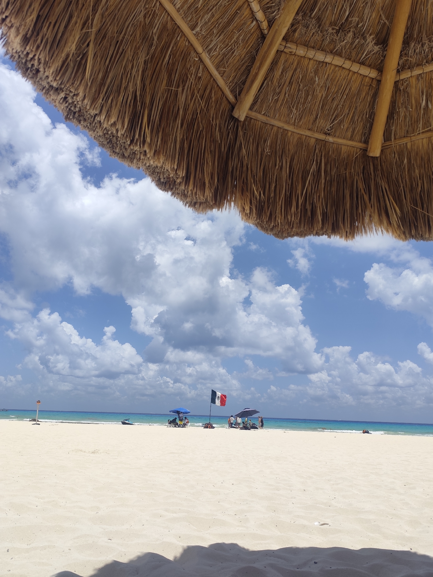 The image depicts a beach scene in Playa del Carmen, Mexico, viewed from under a palapa. The foreground is dominated by white sand, with shadows cast by the palapa. In the mid-ground, there is a view of the turquoise ocean under a partly cloudy sky. A Mexican flag is flying to the right of a blue umbrella. There is a group of people gathered nearby under the umbrella and on the beach. A jet ski is partially visible in the water. A tall orange buoy is placed on the far left of the picture.