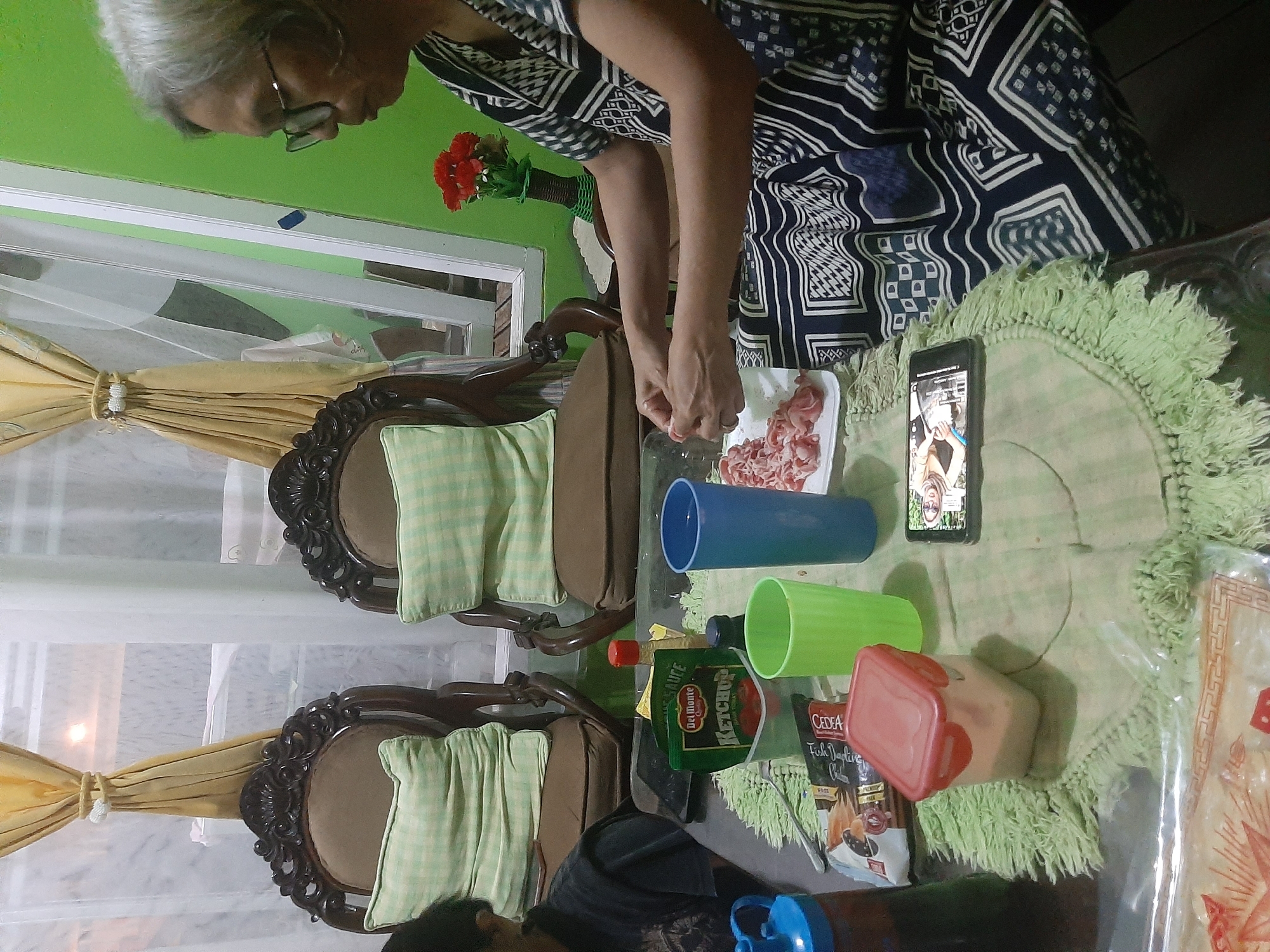 An elderly woman with gray hair and glasses, wearing a dark patterned dress, sits at a table in a room. She is in the process of separating thin slices of raw meat, which are spread on a white rectangular plate in front of her. The table is covered with a light green, fringed tablecloth. To her left, on the table, are various items including a red-lidded plastic container, a green plastic cup, a blue plastic cup, a pouch of Del Monte Ketchup, and a bottle of soy sauce. A smartphone is also visible on the table, displaying an image. In the background, two ornate brown chairs with light green checkered cushions are placed near a window with yellow and white curtains. The wall behind the chairs is painted green. The scene appears to be a home setting in Sumampir, Indonesia, likely a living or dining area.