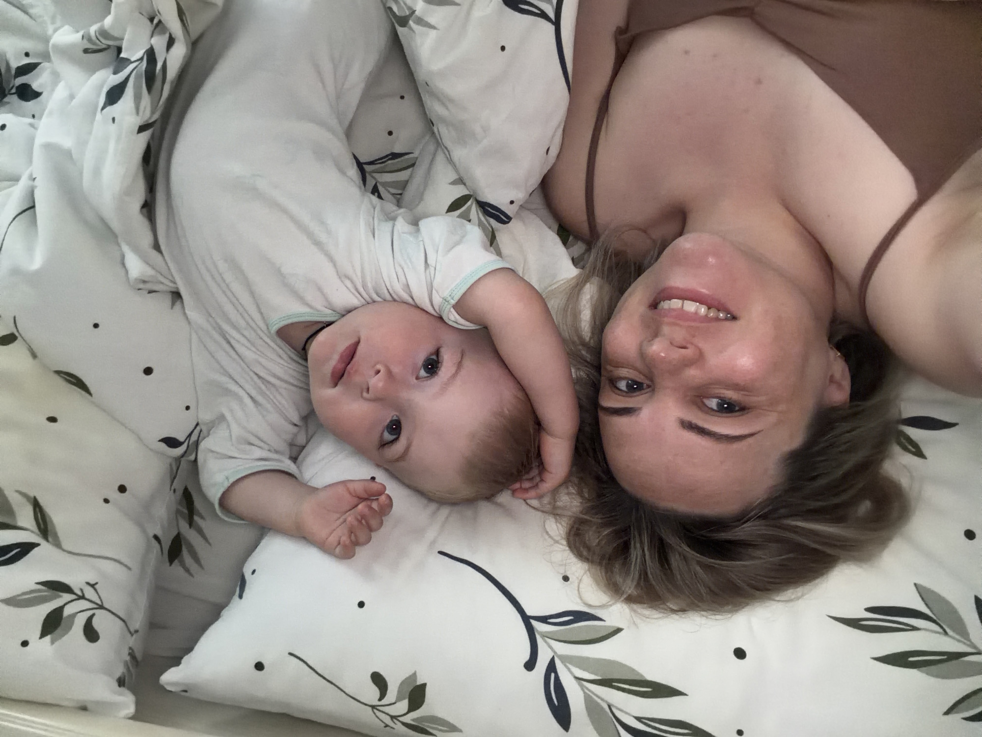 An overhead photograph shows a woman and a baby lying on a bed, both looking directly up at the camera. They are positioned side-by-side on white bedding, which features a pattern of green and dark grey/black leaves along with scattered small dots.
The woman is on the right side of the frame, smiling broadly with her teeth visible. She has light-colored hair and is wearing a brown strap top. Her head is tilted to look at the camera.
To her left, the baby is also looking up at the camera with wide, curious eyes and a slightly open mouth. The baby has light-colored hair and is dressed in a plain, light-colored long-sleeved outfit. One of the baby's arms is bent, with its hand resting near its head.
The scene appears to be an intimate moment indoors, likely in a bedroom. The lighting is soft and even. No specific time of day or outdoor weather conditions are discernible. No text is visible in the image. The location context provided for the image is Votkinsk, Russia.