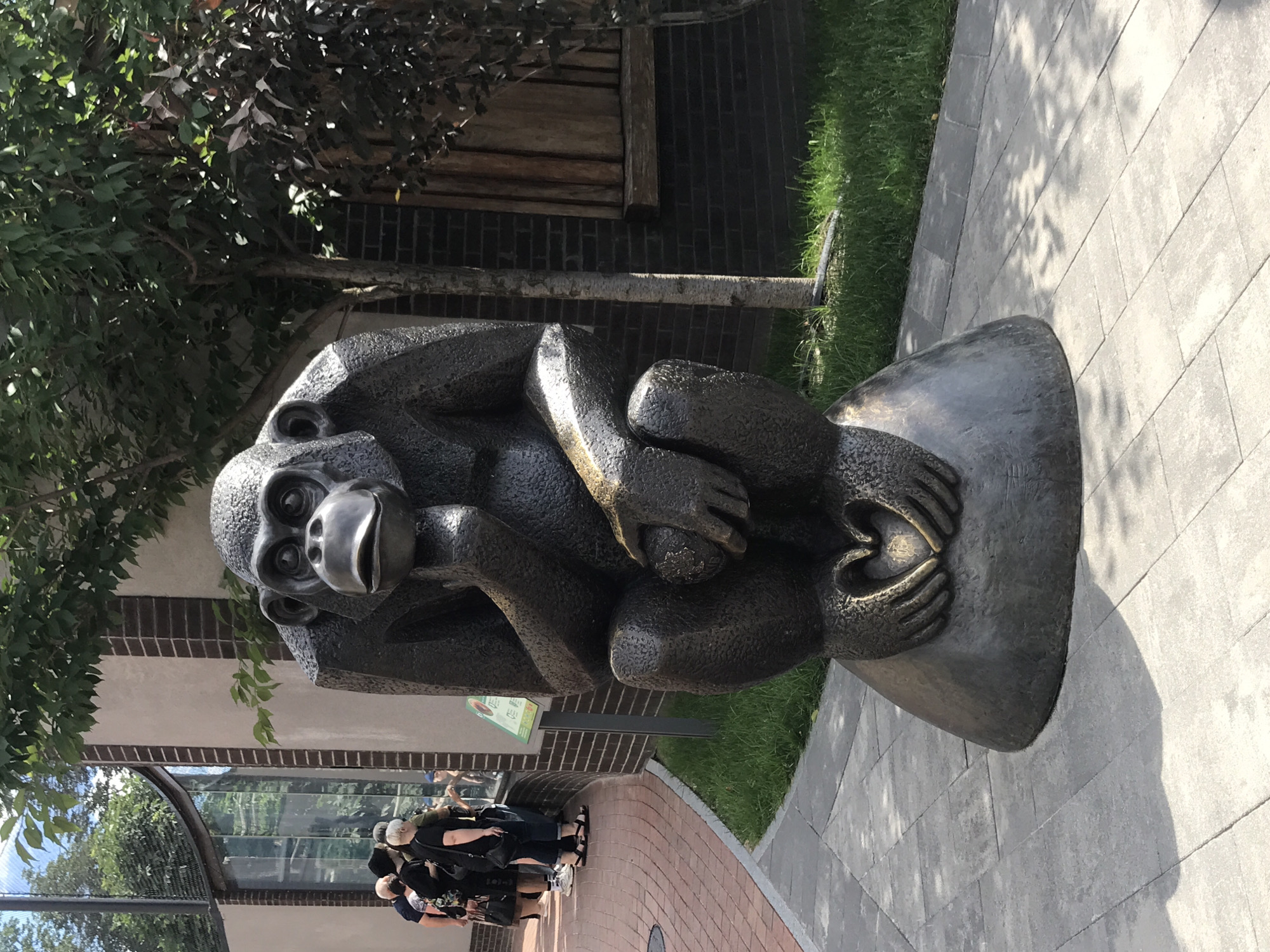 The image shows a bronze sculpture of a chimpanzee sitting with its chin resting on its hand. Its other hand is holding a round object, and its fingers are shaped into a heart. The sculpture is positioned on a rounded base, which sits on a paved walkway. Behind the sculpture, there is a brick building with large windows and trees with green foliage. In the background, several people are visible, walking along a brick pathway and looking into the building. One person is wearing a black hat and black clothing, another is wearing a black top and blue jeans, and a third is wearing a black top and a skirt. A sign with green and white text is also visible near the building. The environment appears to be an outdoor public space, possibly a zoo or a park, in Kharkiv, Ukraine. The lighting suggests it is daytime with shadows cast by the trees.