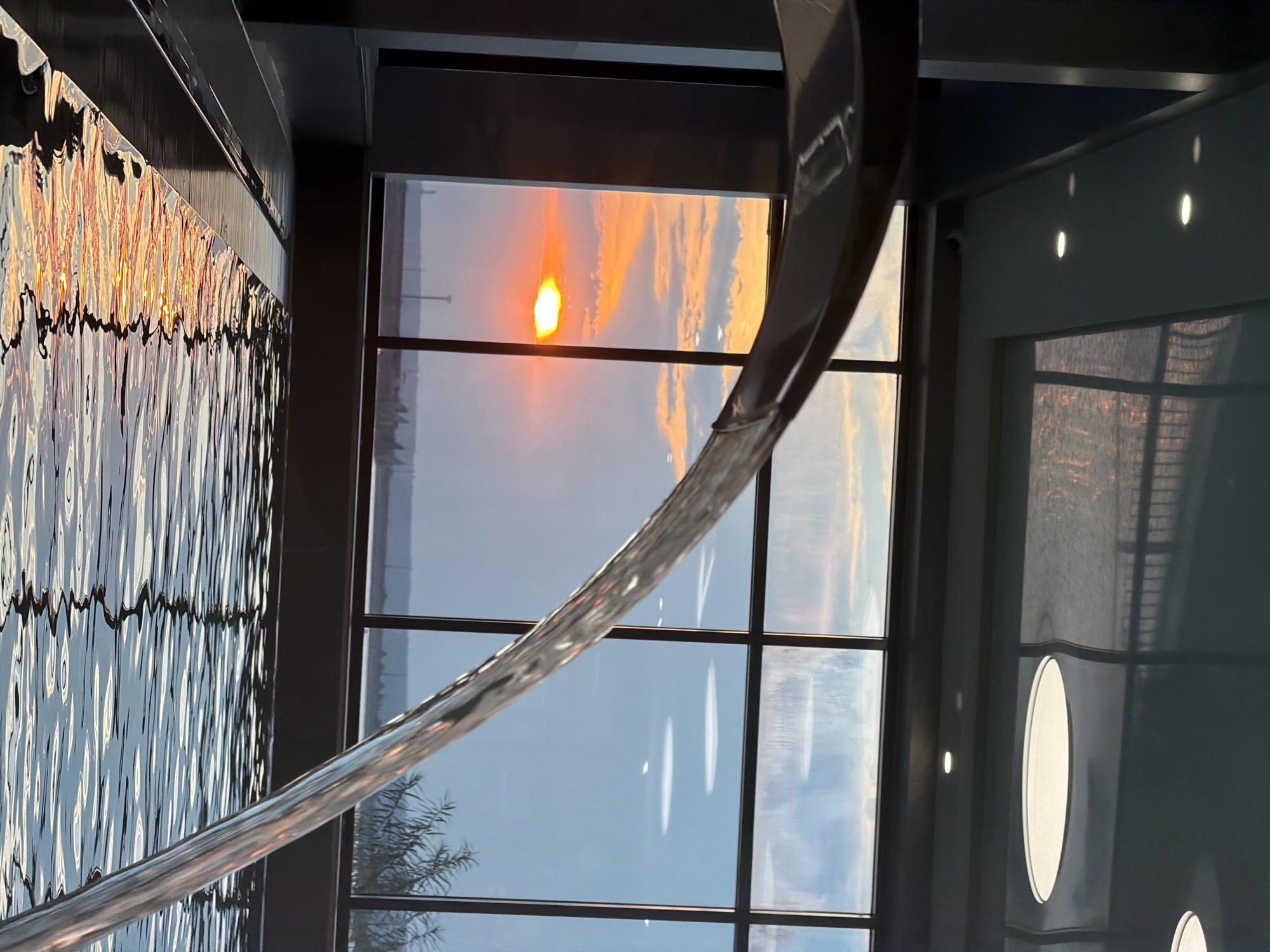 The image captures a serene indoor pool with a large window offering a view of a sunset over Chaikovsky, Russia. In the foreground, a stream of water cascades into the pool, creating ripples that reflect the warm hues of the setting sun. The pool itself is bordered by dark, sleek surfaces, and the ceiling features recessed lighting. Beyond the window, the sky is painted with shades of orange, yellow, and blue, with scattered clouds and the silhouette of distant structures. The overall impression is one of tranquility and natural beauty observed from a modern, well-appointed indoor space.