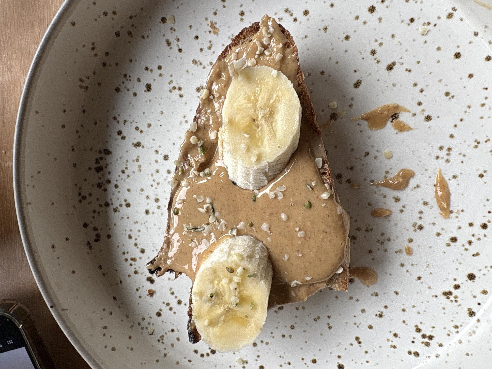 A close-up, high-angle shot depicts a slice of toast on a speckled plate. The toast is triangular and spread with a tan-colored nut butter, possibly peanut or almond. Two slices of banana are placed on top of the spread. Scattered seeds, likely hemp, are sprinkled across the toast. Stray smears of the nut butter are on the plate next to the toast. A small portion of a smartphone or electronic device is visible in the lower left corner of the frame. The background is a light-colored wooden surface. The plate itself is off-white with brown speckles. There is no indication of the location being in London, United Kingdom.