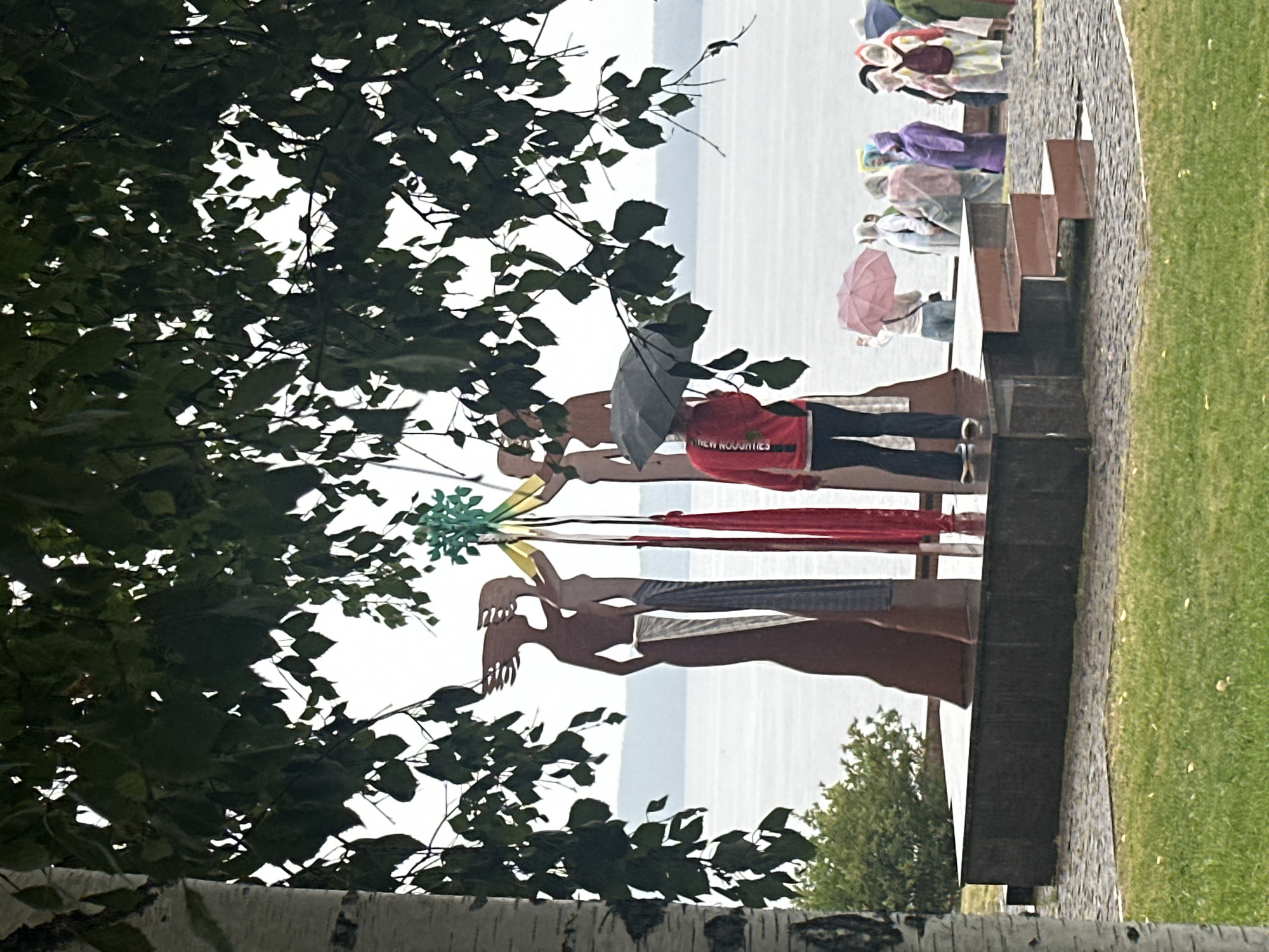 The image shows a scene in Petrozavodsk, Russia, featuring a sculpture of four stylized female figures on a tiered platform near a body of water. A person holding a black umbrella stands in front of the sculpture. In the background, a group of people are visible, many of them holding umbrellas or wearing raincoats, suggesting inclement weather. The sculpture is partially obscured by the branches and leaves of a large tree in the foreground. The environment includes a grassy area, a paved walkway, and the water with a distant shoreline.