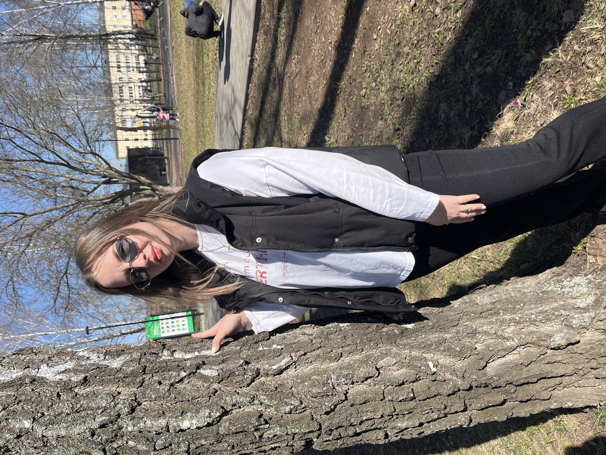A young woman wearing sunglasses, a white shirt, a black vest, and black pants is posing outdoors. She is leaning against a tree trunk with her hand resting on the bark. In the background, there is a park-like setting with bare trees and a building in the distance. A park information sign is also visible. The location is Zav'yalovo, Russia.