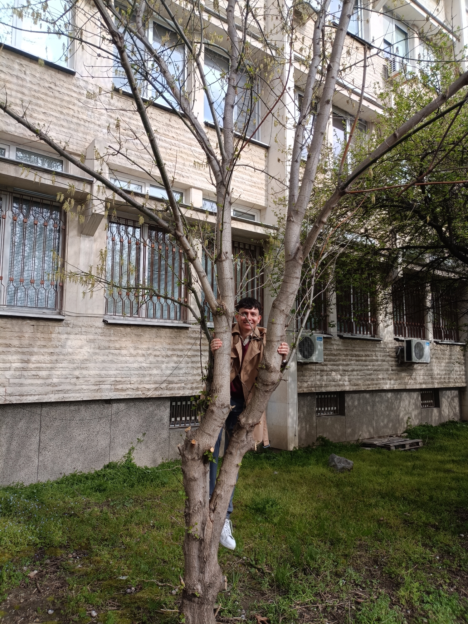 A person stands behind a tree on a grassy lawn in Plovdiv, Bulgaria. The person has short dark hair, is wearing a brown overcoat, jeans, and white shoes. The person is holding onto the tree, and smiling at the camera. Behind them is a light-colored, multi-story building with barred windows and air conditioning units. The building is likely located in an urban environment. There is another tree to the right of the building and the person.