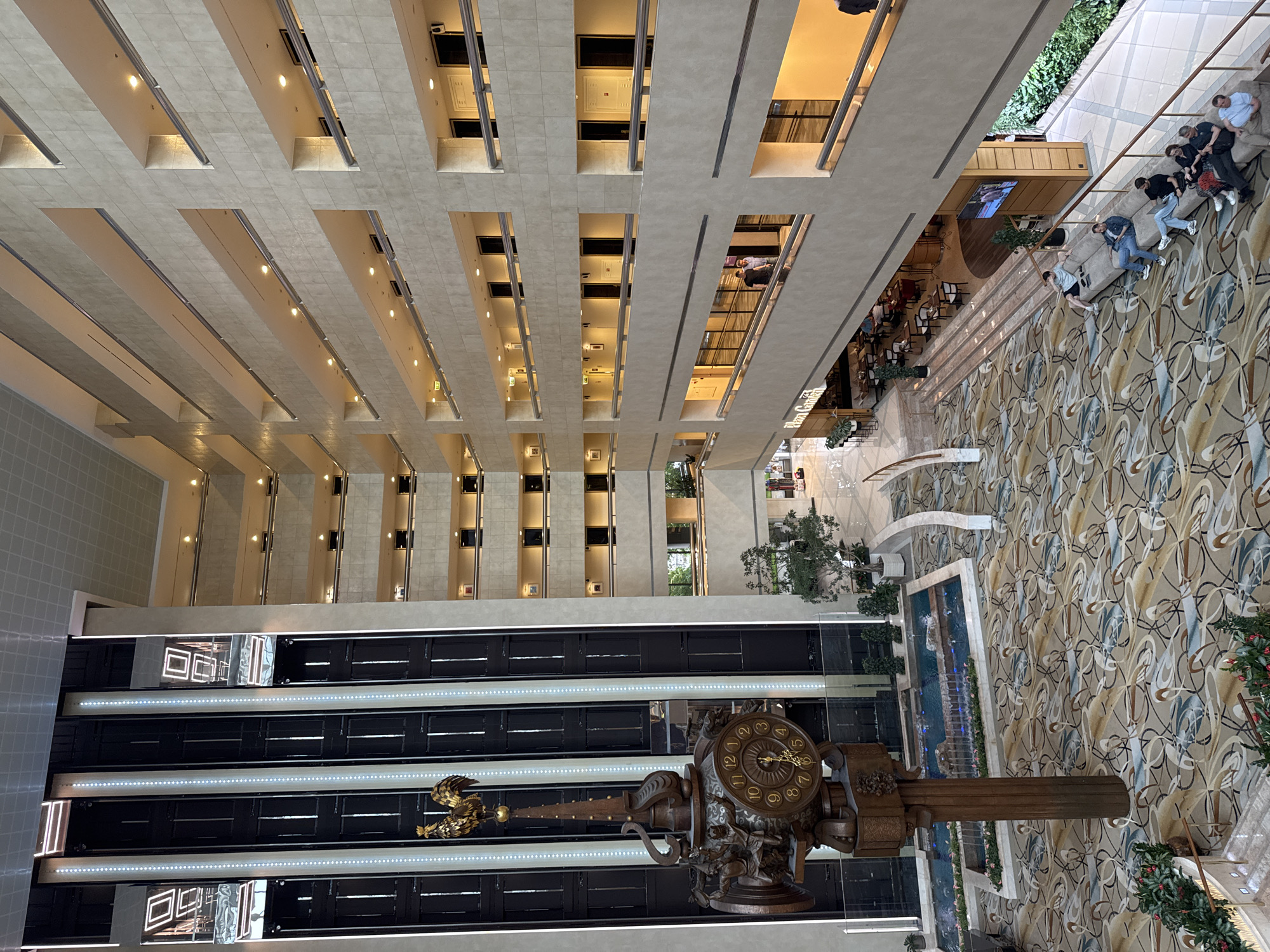 The image shows the interior of a multi-story building with a large open atrium. The view is from an upper level, looking down at the lobby. The lobby features a large, ornate clock sculpture, a water feature, and a patterned carpet. Several people are visible on a balcony overlooking the lobby. The building has a grid-like architectural design with multiple floors and recessed windows.
Based on the image's characteristics and the presence of a large clock sculpture, it is plausible the location is the Radisson Collection Hotel in Moscow, Russia.