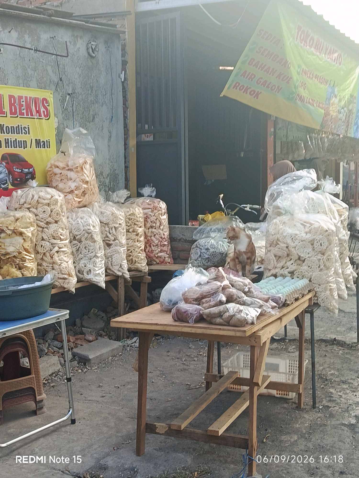 The image depicts an outdoor market stall in Keboansikep, Indonesia, bathed in the harsh light of late afternoon. A weathered, concrete wall forms the backdrop, marked by exposed conduits and what appear to be remnants of older structures. To the left, a faded yellow banner with bold red lettering announces "BEKAS" and "Kondisi Hidup / Mati", likely indicating a sale of second-hand items or perhaps a dire warning. Below this, a red car is depicted.
Dominating the foreground is a wooden table laden with various goods. Piled high in clear plastic bags are numerous types of crackers and snacks, some with intricate, circular patterns, others with more irregular shapes, in shades of beige, white, and reddish-brown. A green plastic basin sits on a smaller, blue-topped table to the left, filled with more snacks. A ginger and white cat is perched on the wooden table, looking downwards, surrounded by bags of snacks and an assortment of packaged food items, including what appear to be eggs laid out in a pale green tray. Further to the right, more plastic bags of snacks are visible, some resting on wooden planks. The ground is a mix of concrete and dirt, with scattered debris.
Above the stall, a green banner with yellow lettering reads "TOKO RUKU YAKTO" and lists various items for sale, including "SEDIA : SEMBAKO" (groceries), "BAHAN KUE" (baking ingredients), "AIR GALON" (water gallons), and "ROKOK" (cigarettes), alongside "ANEKA PLASTIK" (various plastics) and "LPG". The overall impression is one of a humble, local market. The timestamp "06/09/2026 16:18" is visible in the lower right corner, along with "REDMI Note 15" on the lower left.