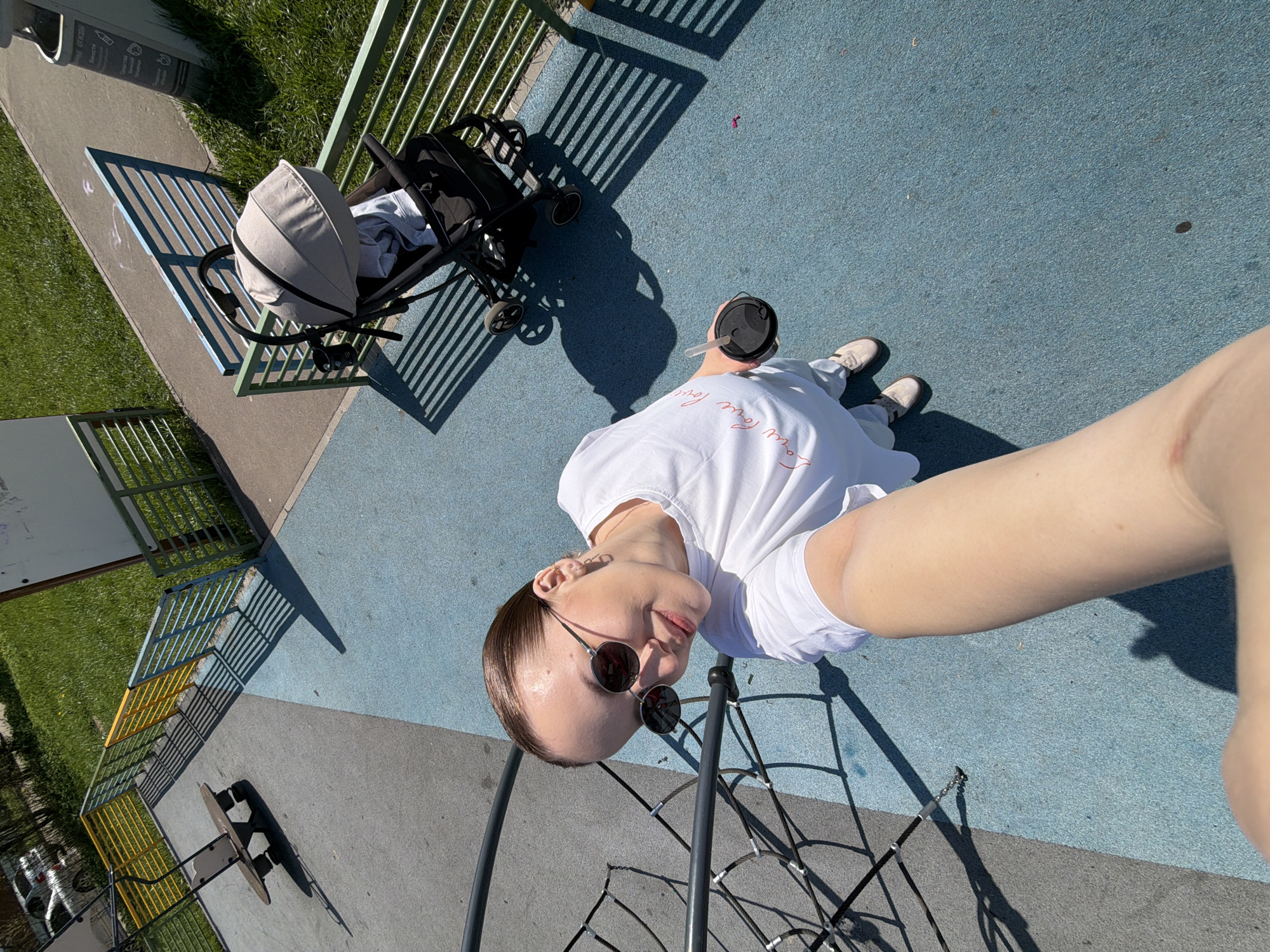 The image appears to be a selfie taken outdoors in a playground or park, potentially in Reshetnikovo, Russia, though this cannot be confirmed without further information. A person wearing sunglasses and a white t-shirt is holding a drink, perhaps coffee or a smoothie, with a straw. They are standing on a blue surface, possibly a playground floor. In the background, there is a stroller parked near a green metal fence. The area includes grass, sidewalks, and playground equipment, with a climbing structure visible in the foreground. The lighting suggests it is a sunny day.