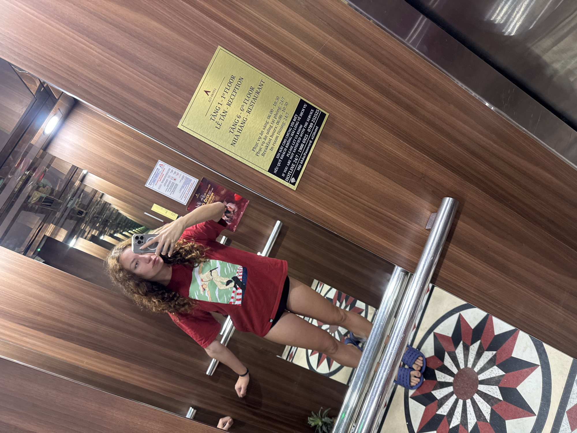 In a hotel elevator in Hanoi, Vietnam, a young woman with curly brown hair takes a selfie in the mirror. She is wearing a red t-shirt with a graphic print and black underwear. Her left arm is resting on the elevator wall, and her right hand is holding a smartphone. The elevator walls are made of wood-paneled material, and there is a mirrored surface reflecting the scene. A sign on the wall indicates "A25 HOTEL" and provides information about floor levels and services, including reception hours and in-room dining. The elevator floor has a circular, patterned mat with red, black, and white geometric designs. Blue flip-flops are visible on the woman's feet.