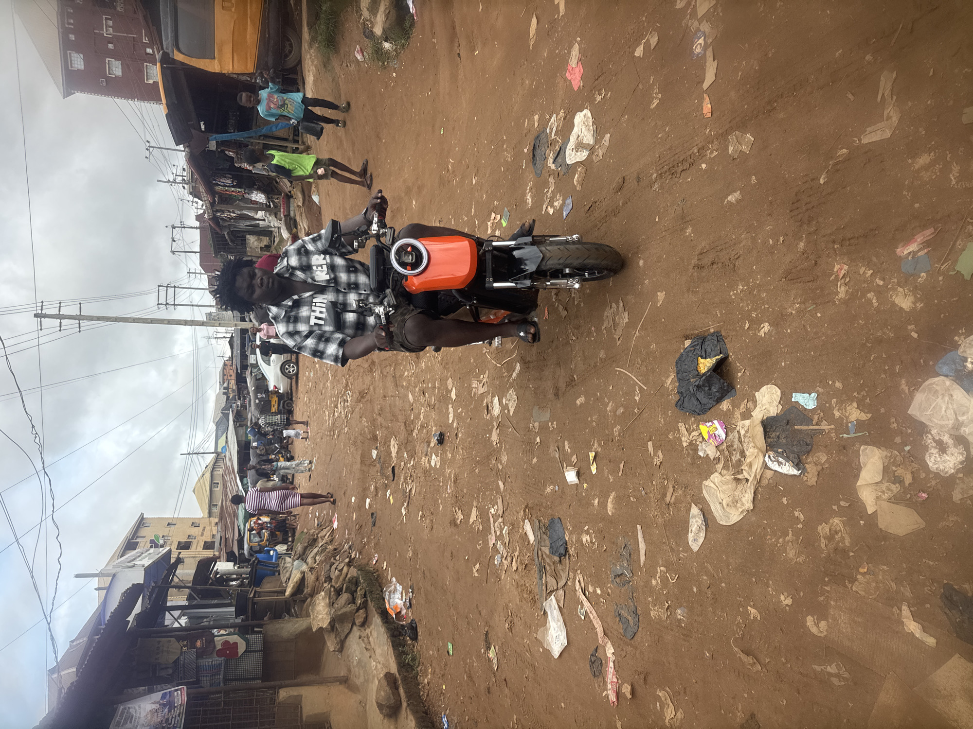 A medium shot captures a person on a black and orange electric scooter on a dirt road in Nkpor, Nigeria. The individual, wearing a checkered shirt and dark shorts, is looking directly at the camera. The road is littered with debris, including plastic bags, paper, and other refuse. Buildings line the sides of the road, appearing to be shops and residences. In the background, several other people, including children, are visible. Overhead, power lines crisscross the sky, and the weather appears overcast. A yellow vehicle is parked to the right of the frame. The overall impression is one of a busy, perhaps impoverished, urban environment.