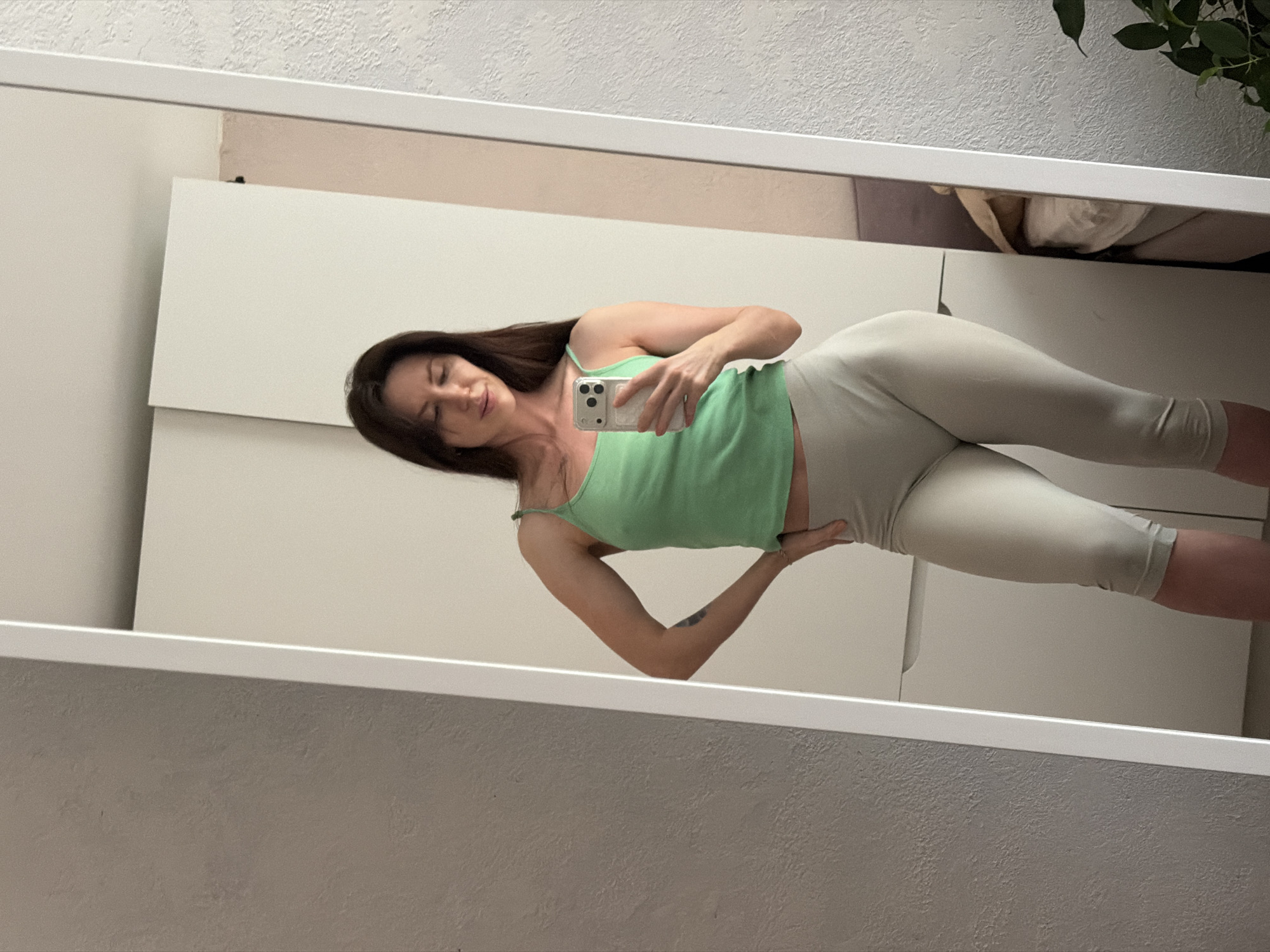 A woman with long brown hair is taking a selfie in front of a mirror. She is wearing a light green tank top and light gray leggings. She is holding a white smartphone in her right hand. The background consists of a white closet or wardrobe and a textured gray wall. A plant is visible in the upper right corner. The location in the city of Perm, Russia cannot be determined from the image.