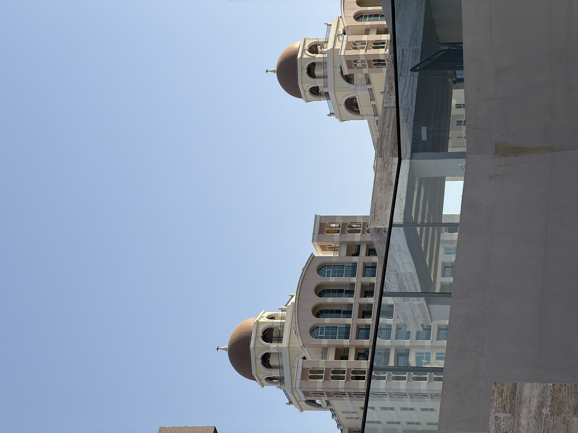 The image captures an upward view of modern buildings with distinctive architectural elements under a clear, bright blue sky. In the midground, a light-colored, multi-story building featuring a large, dark brown dome is prominent. This building showcases numerous arched windows and balconies, suggesting a classical or Mediterranean design influence. Another similar building, also topped with a dome, is partially visible further back.
A significant glass barrier or window stretches across the foreground, reflecting portions of the buildings and the sky, creating an interesting interplay of light and imagery. Through the glass, glimpses of interior structures and a dark handrail are discernible. To the right, a solid, light gray concrete or stucco wall occupies a large section of the foreground, displaying some minor discoloration or staining. The bright sunlight indicates it is daytime with clear weather. No people or specific activities are depicted, and there is no visible text.