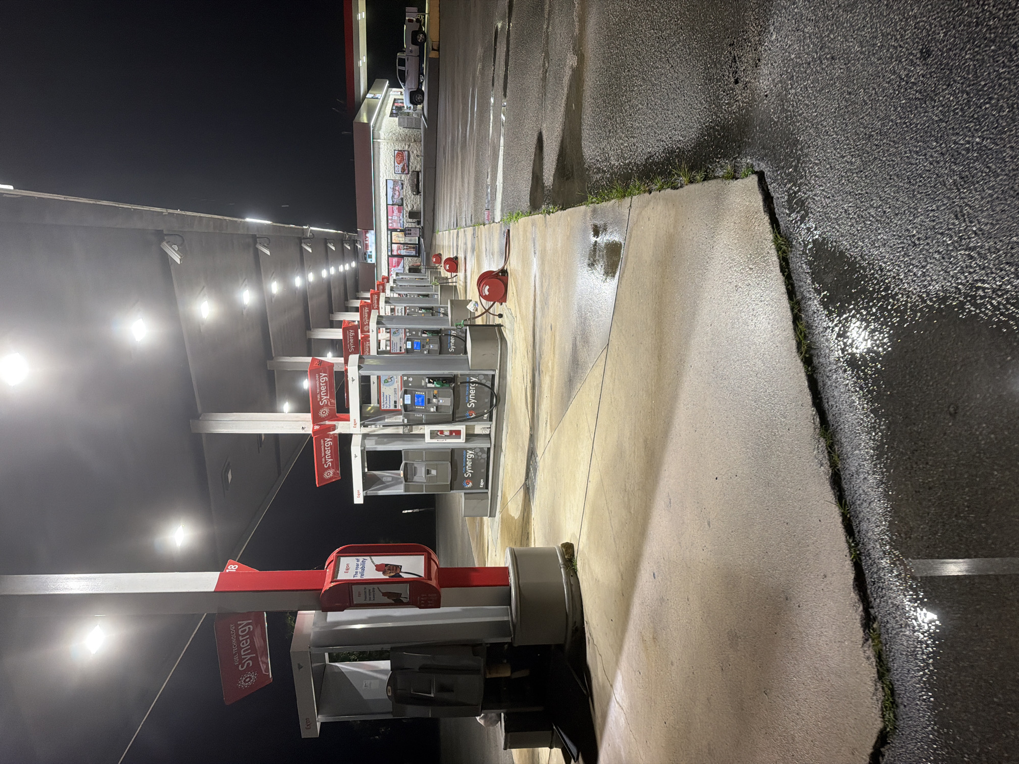 The image shows a gas station at night in Wildwood, United States. The gas station features multiple pumps under a lit canopy. Several red hoses with reels are visible on the ground near the pumps. The ground is wet, with puddles reflecting the overhead lights. In the background, a convenience store is visible with lights on inside and a white pickup truck parked in front of it. The scene appears to be deserted.