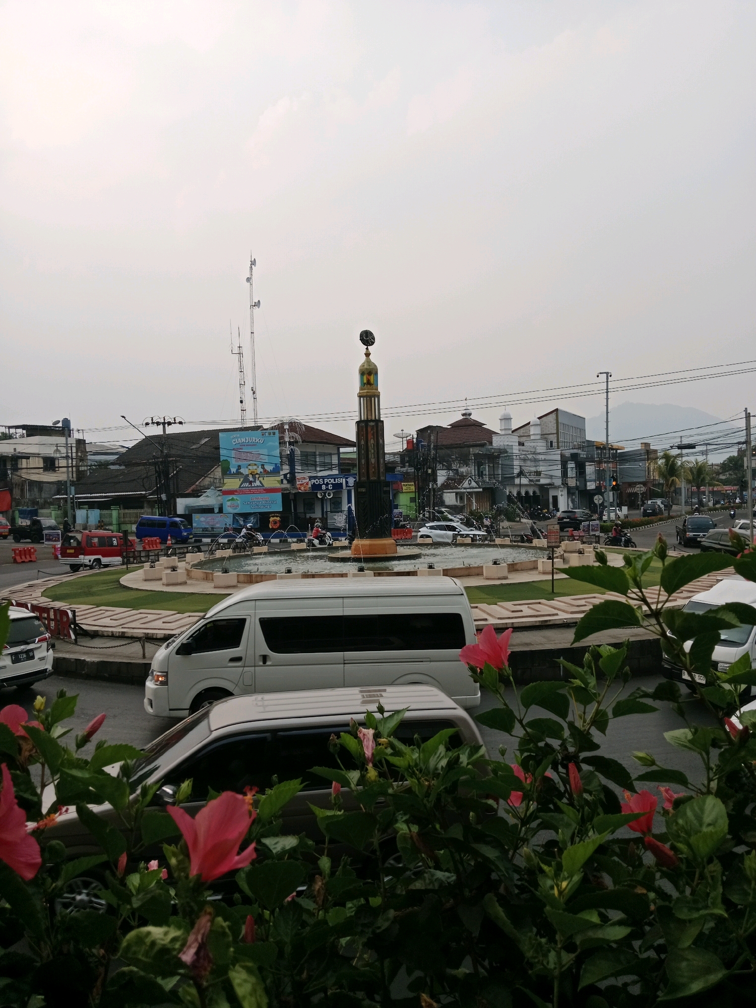The image captures a street view in Cianjur, Indonesia, centered around a roundabout with a monument. In the foreground, vibrant pink hibiscus flowers and lush green foliage are prominent. Beyond the flowers, a white van is parked on the street, and part of another white van is visible to its right. The roundabout features a fountain and a tall, cylindrical monument topped with a golden cap. Surrounding the roundabout are various buildings, including shops and businesses, some with advertisements on their facades. A radio tower stands tall in the background, and distant mountains are visible under an overcast sky. Several vehicles, including cars and motorcycles, are present on the street, indicating traffic activity. A "POS POLISI" (Police Post) sign is visible on one of the buildings. The scene depicts a typical urban environment with traffic, commercial activity, and a public monument.