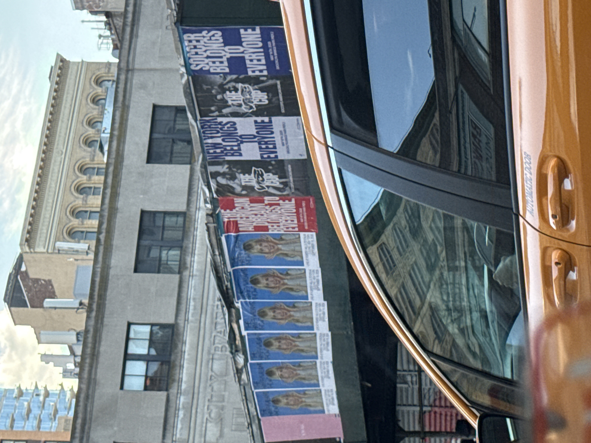 The image shows a view of a building facade adorned with posters and a portion of a yellow vehicle, likely a taxi, in New York City, United States. The building is of typical urban architecture, with rectangular windows and detailed cornices. The posters include messages like "Soccer Belongs to Everyone" and "New York Belongs to Everyone," along with images of people. One set of posters features a woman with blond hair and a light-colored top. The posters are attached to the building's exterior, suggesting some event or campaign. The side of the vehicle has "AUTOMATIC DOOR" written on it.