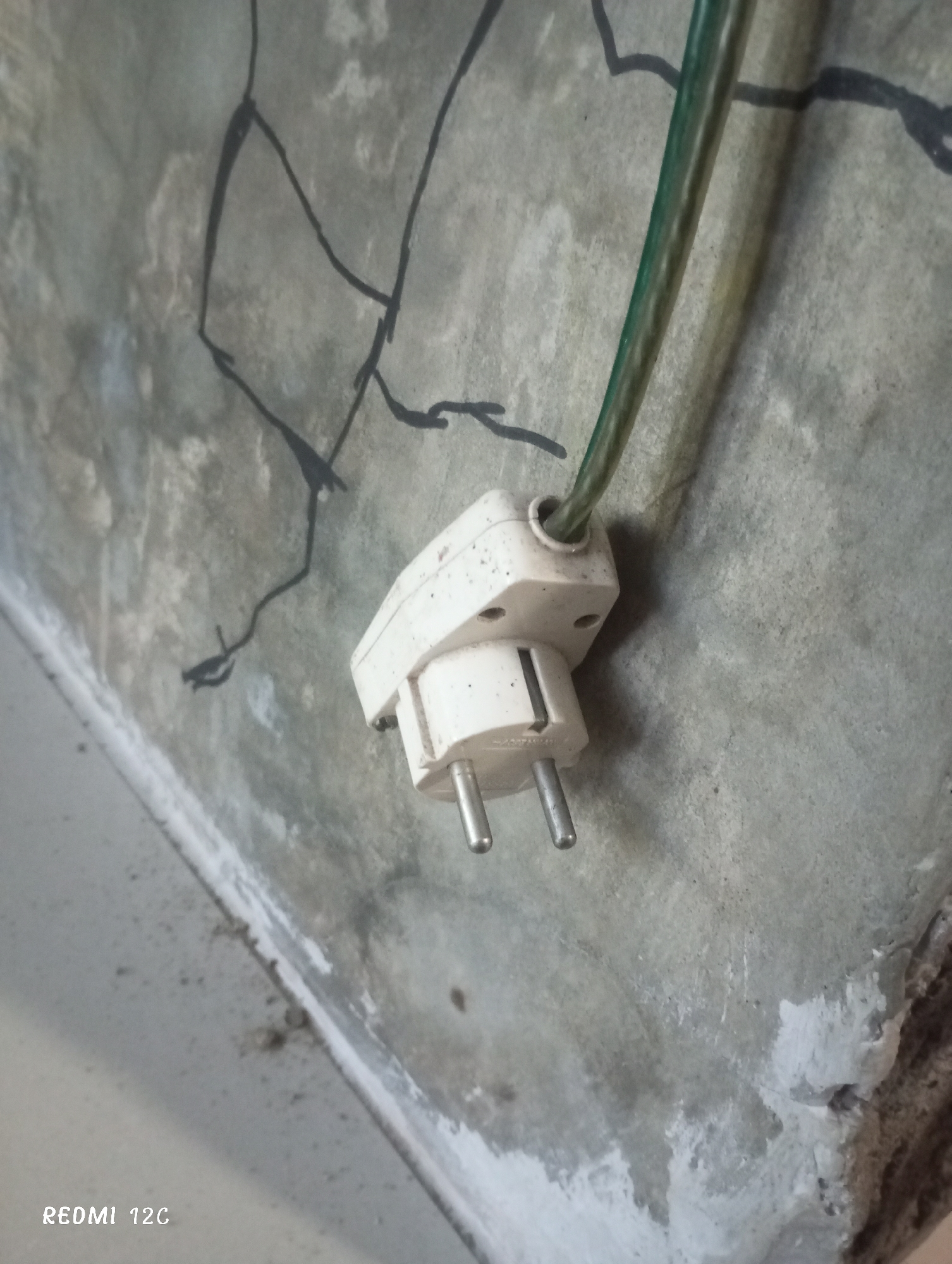 A European plug is emerging from a wall. A green cord leads into the top of the plug. The wall is made of grey concrete and marked with black lines. The bottom left corner of the photo contains the watermark "Redmi 12C," indicating the photo was taken with that type of phone. The photo appears to be taken at an oblique angle. Location in Lhokseumawe, Indonesia, is not independently confirmable.
