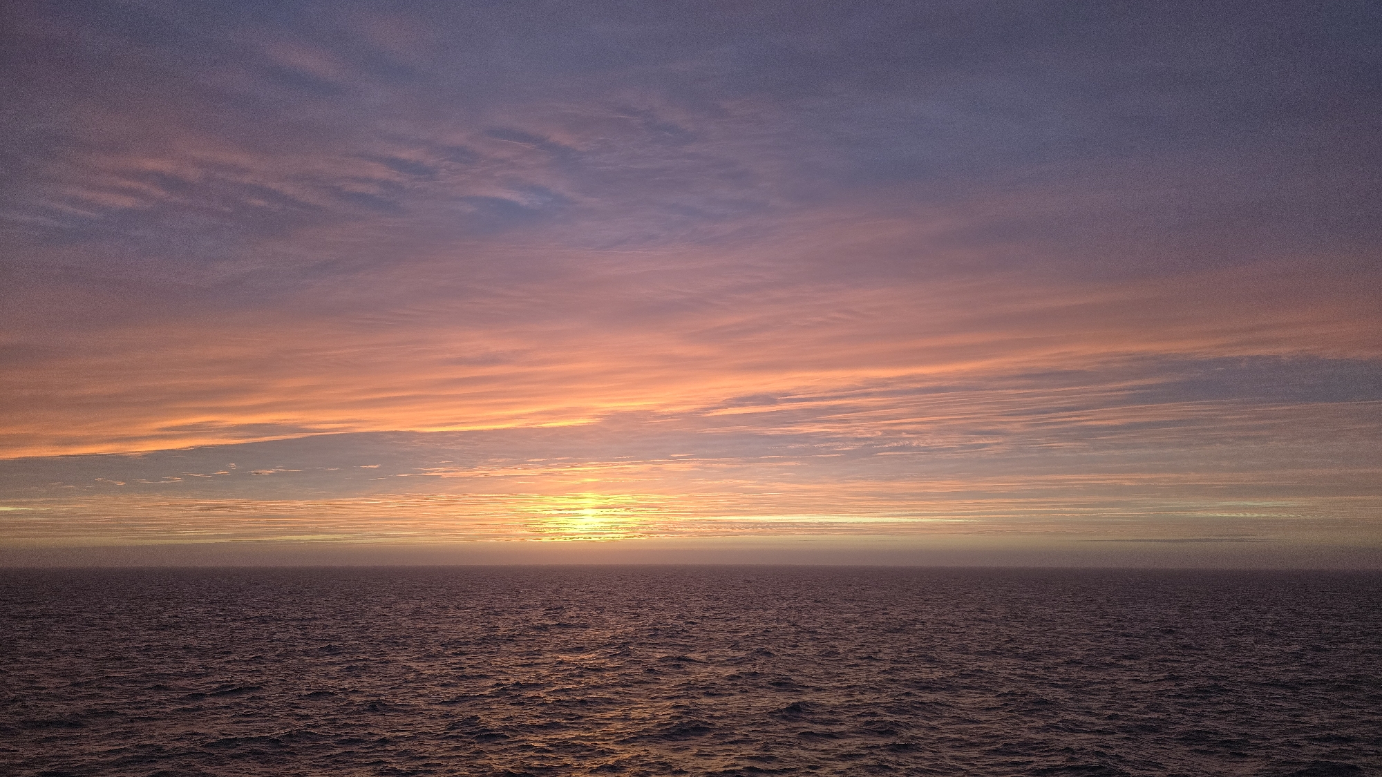 A photograph captures the sunset over the ocean. The sky is a mix of pink, orange, and purple hues, with long, horizontal clouds stretching across the frame. The sun is setting on the horizon, casting a golden glow on the water's surface. The ocean is dark with waves. The location is not specified.