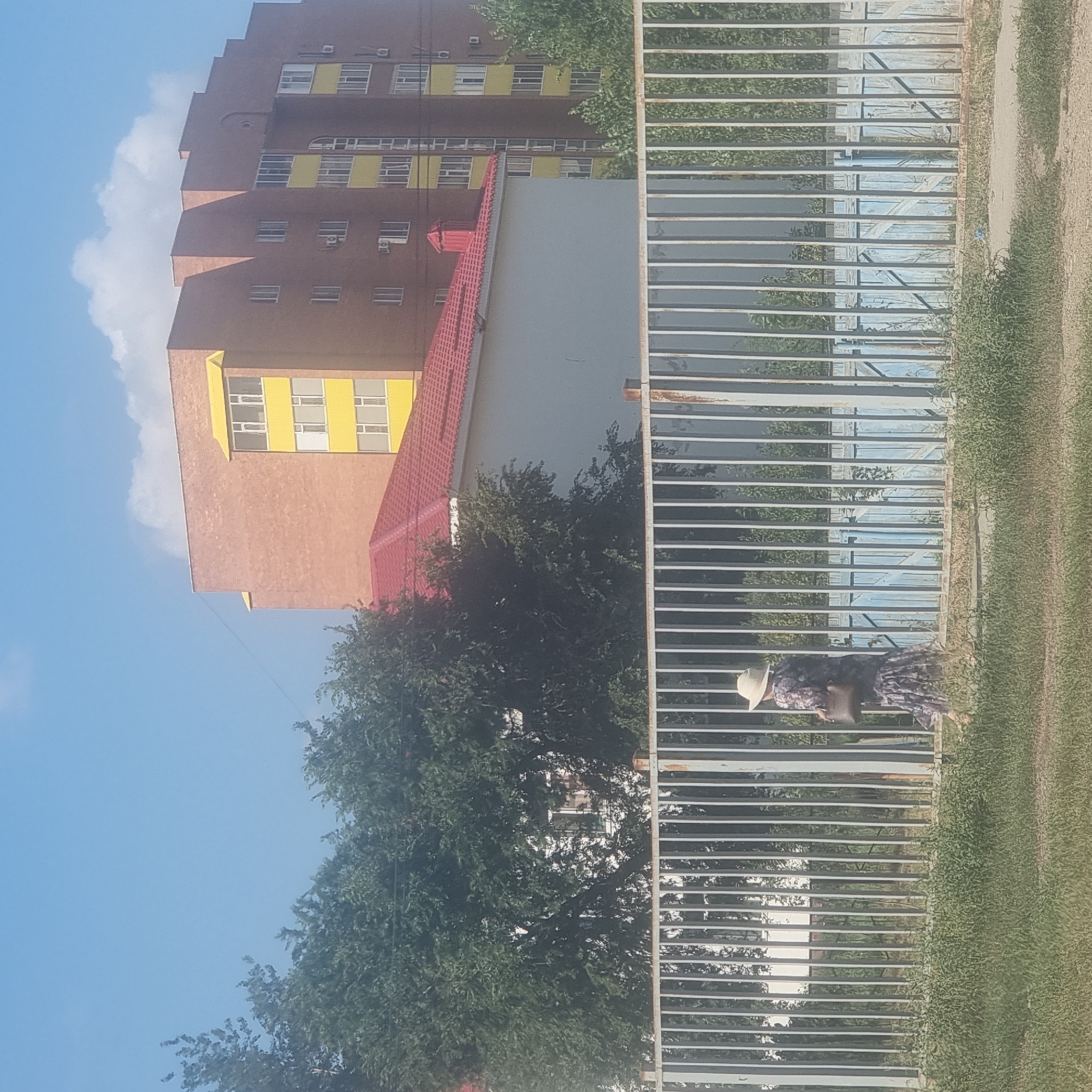 The image depicts a street scene in Aktobe, Kazakhstan on a sunny day with a clear blue sky and scattered clouds. In the foreground, a person wearing a floral dress and a straw hat walks past a metal fence. The person is carrying a brown purse. Behind the fence, there is a building with a red roof and trees. Further in the background, a multi-story brick building with yellow accents is visible. The overall impression is of an urban environment.