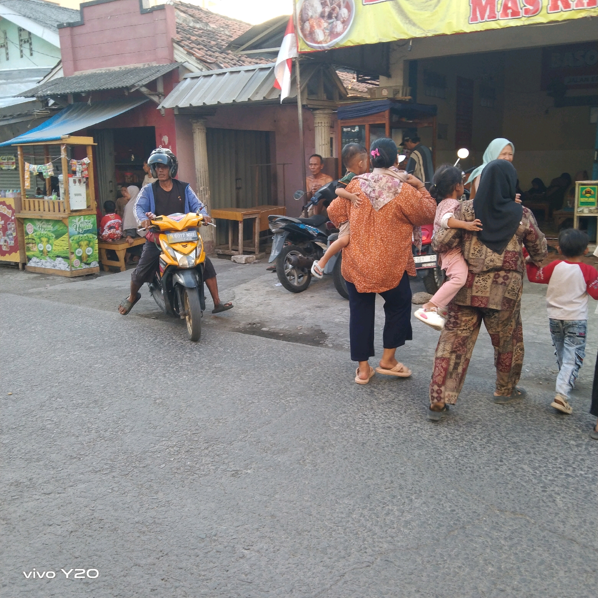 The image depicts a street scene in Karawang, Indonesia, likely during daylight hours given the ambient lighting. A man wearing a blue jacket, black t-shirt, and dark pants is riding a yellow motorcycle with a black front fender and a license plate that reads "B 6017 MUF". He is wearing a black helmet. The motorcycle is positioned in the foreground and appears to be moving or stopped on an asphalt road.
To the right of the motorcycle, a woman in a patterned orange and white top and dark pants is carrying a young child on her back. The child is wearing pink pants and white shoes. Another woman, wearing a black hijab and a patterned brown, red, and beige outfit, is also present, holding the child. To the far right, a young boy in a red and white shirt and blue jeans is walking.
In the background, there are several buildings with tiled roofs. One building has a red facade and a sign with Indonesian text, possibly a business name or advertisement. There is a wooden stall or kiosk on the left side of the image with products displayed. Other individuals are visible in the background, seated on wooden benches. Two motorcycles are parked behind the people in the foreground. The overall environment suggests a commercial or residential area within a city.
