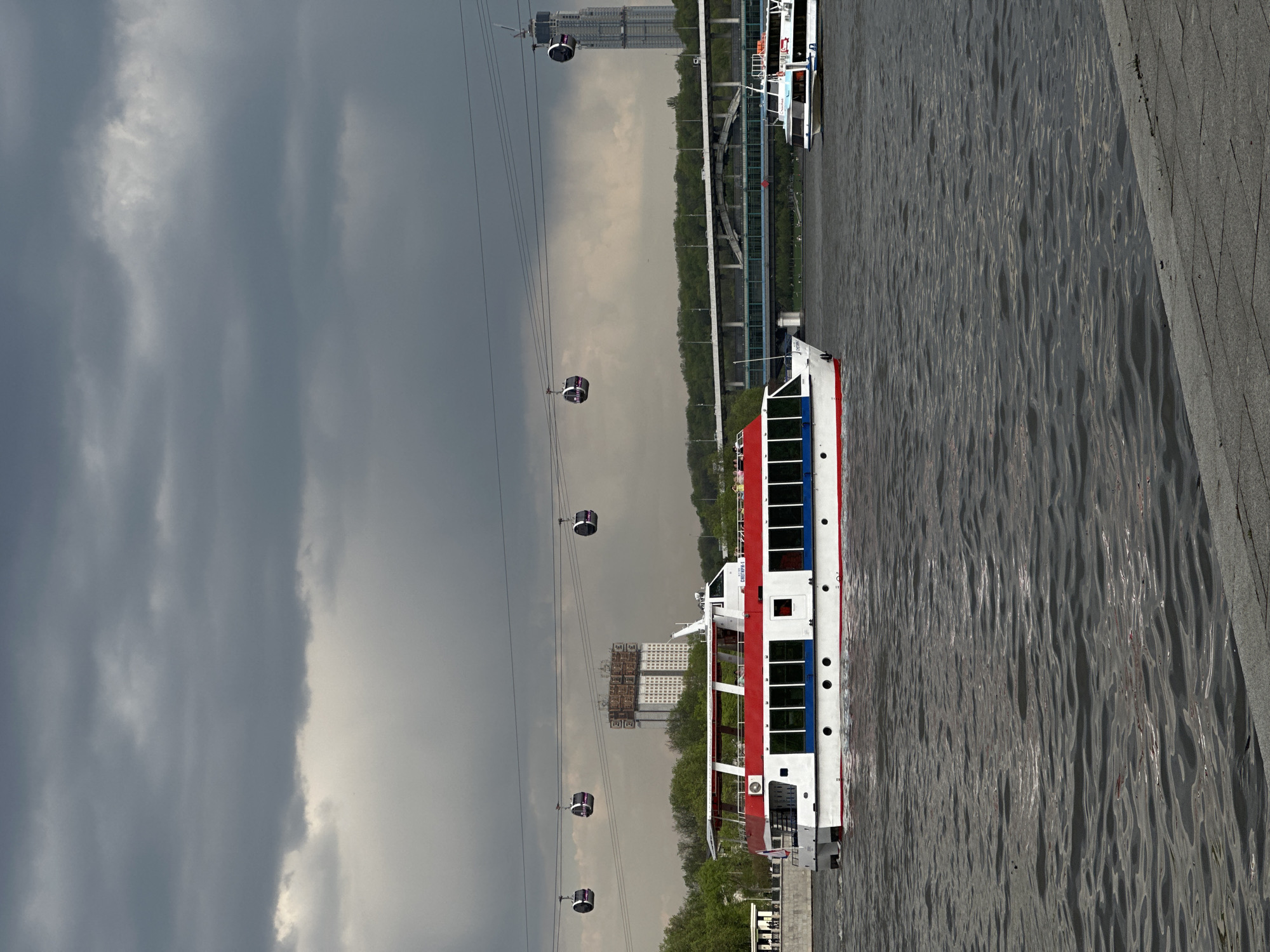 The image depicts a scene in Moscow, Russia, featuring a river, a boat, and a cable car system. The boat is red, white, and blue, and is docked near a concrete embankment. The cable car system has several cars suspended on cables stretching across the river. In the background, buildings and trees are visible. The sky is overcast with dark, stormy clouds.