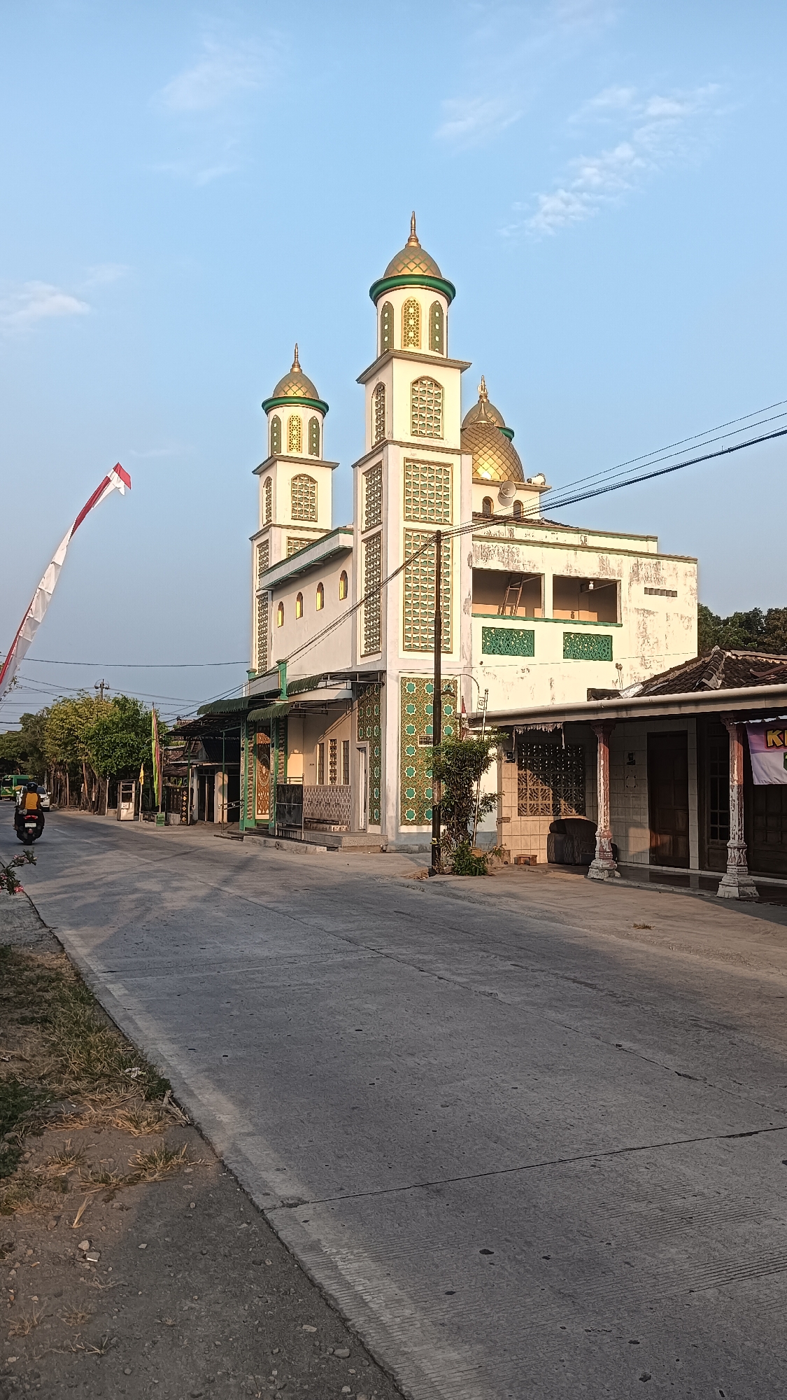 The image depicts a mosque in Kedungupit, Indonesia, on a clear day with a blue sky. The mosque features a prominent white facade with gold domes and green accents on its minarets and decorative patterns. A red and white Indonesian flag flutters on a pole to the left of the mosque. The mosque is situated beside a paved road, with a few trees and other buildings lining the street. A person is seen riding a yellow scooter in the distance on the left side of the road. The overall scene captures a tranquil street view of a religious building in a village setting.
