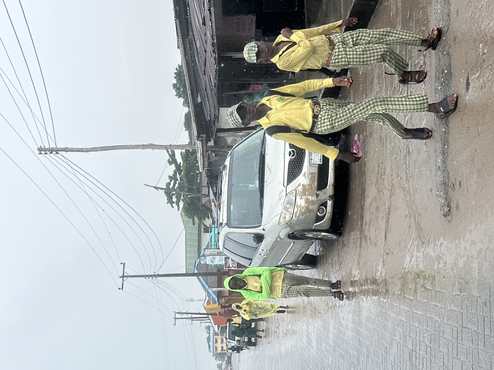 This is a vertical, eye-level shot of a street scene in Badagry, Nigeria. The primary subjects are several young people, appearing to be boys, dressed in matching yellow shirts and plaid pants. Two of them are standing prominently near a silver car parked on the side of a wet, paved road. They are wearing dark sandals and appear to be posing or waiting. Another individual in a bright green hooded jacket and similar plaid pants is also visible further down the street. In the background, there are buildings with corrugated metal roofs, more cars, and utility poles with numerous wires. The ground is wet and reflects the overcast sky, suggesting it is raining or has recently rained. The overall mood is somewhat subdued, possibly due to the weather. There is some visible text on the license plate of the silver car, which appears to read "110 LAG." The time of day is difficult to determine precisely due to the overcast conditions, but it seems to be daytime.