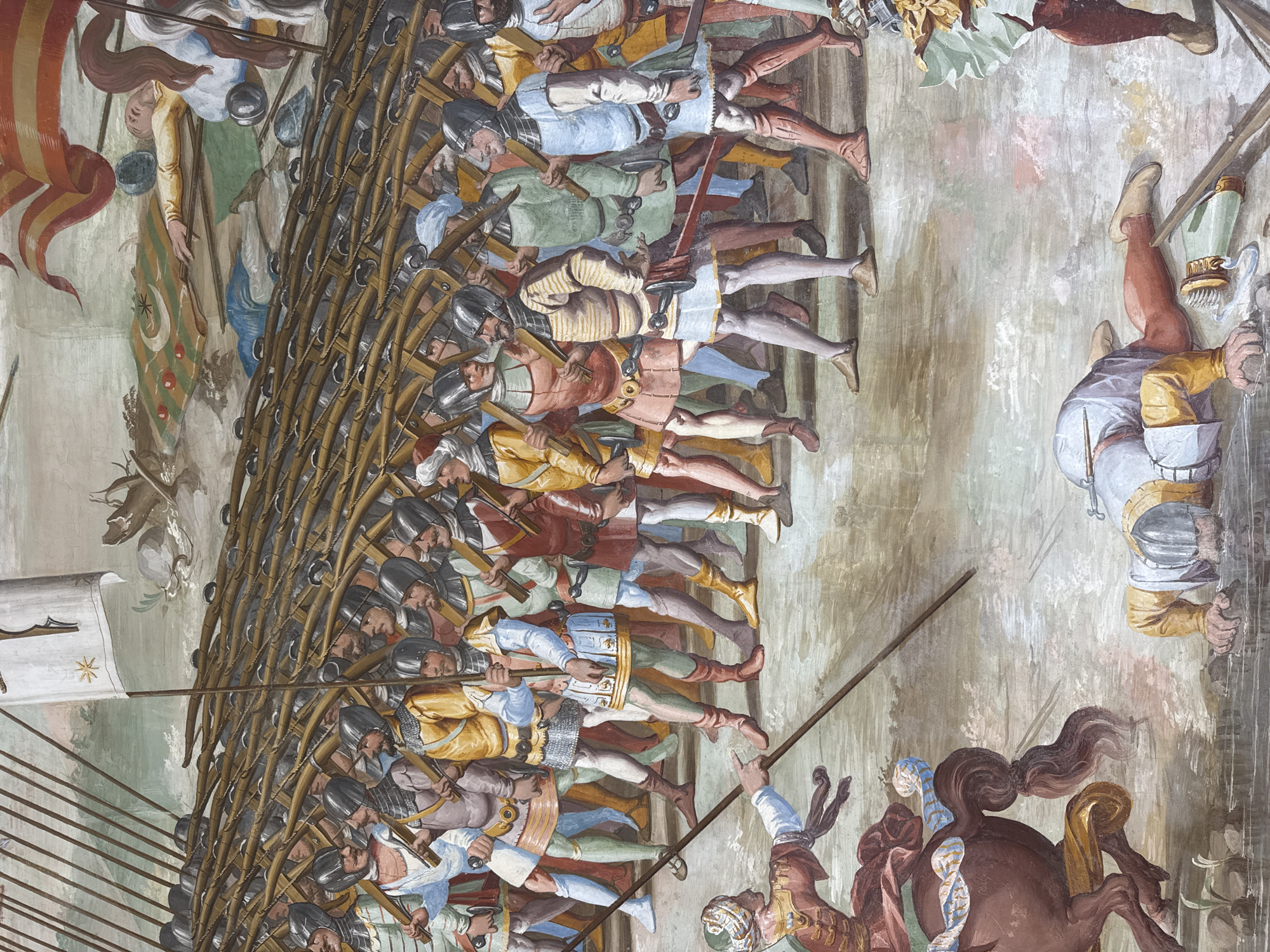 This is a fresco, likely part of a larger narrative mural, depicting a battle scene. A large formation of soldiers, armed with spears and wearing armor and helmets, are arranged in a disciplined line. Their clothing is varied in color, including reds, yellows, blues, and greens, suggesting different ranks or factions. The soldiers' faces show a range of expressions, from focused determination to what appears to be exhaustion.
In the foreground, a horse is depicted, with a figure on its back, possibly a commander or a warrior. The horse has a flowing mane and tail, and is colored a deep brown. The overall scene suggests a chaotic but organized military engagement. The fresco style, with its visible brushstrokes and muted color palette, gives the artwork a sense of age and historical significance. The background is rendered in soft, earthy tones, with hints of what might be a rocky or barren landscape. There is no visible text in the image. The lighting appears to be diffuse, suggesting it's either indoors or a scene painted without a strong emphasis on a specific time of day. The overall impression is one of historical reenactment or a depiction of a past conflict.