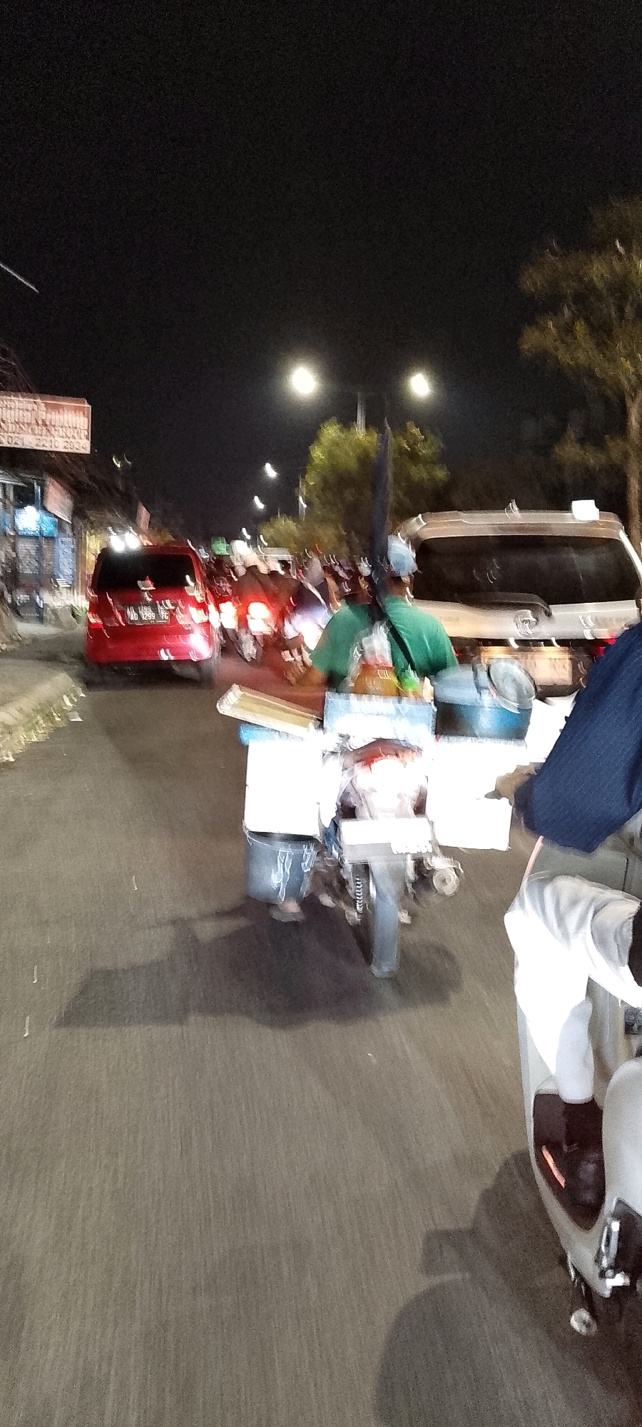 The image captures a nighttime street scene in Bekasi, Indonesia, likely a busy commercial area. The foreground shows the right side of a scooter, with its driver partially visible. The scooter is carrying a cart laden with various items, including what appear to be bottles, containers, and possibly food supplies. Behind the scooter, a person wearing a green shirt and a cap is seated, with an umbrella strapped to their back. Further down the road, a red car is stopped, and other vehicles and motorcycles are in motion, indicated by motion blur. Streetlights and vehicle headlights illuminate the scene, casting shadows on the asphalt road. Buildings with signage are visible on the left side of the street. The overall atmosphere suggests a typical urban environment at night, possibly during a traffic jam or at a marketplace.