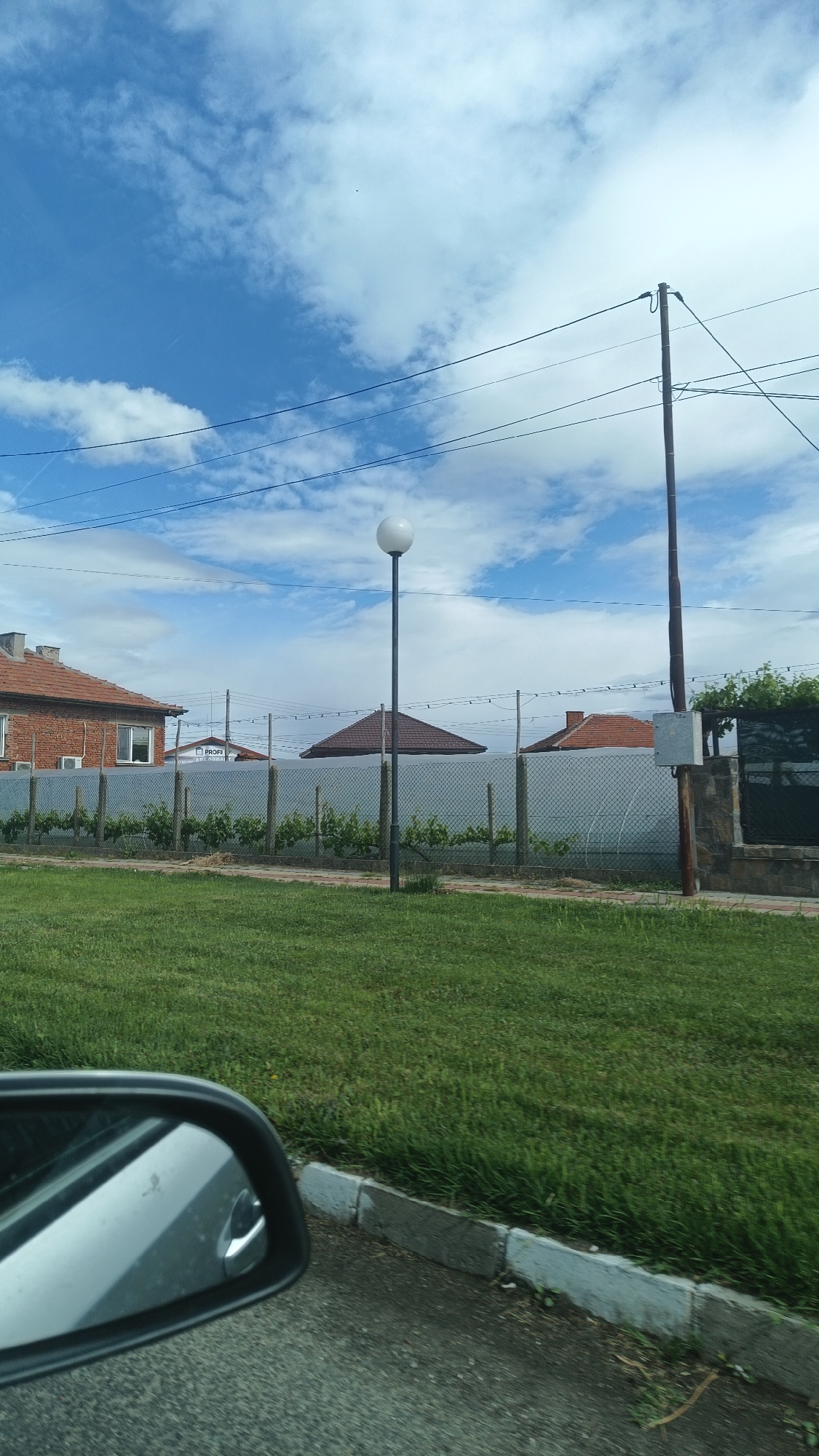The image depicts a scene in Popovitsa, Bulgaria, captured from inside a moving vehicle, as evidenced by the side mirror visible in the foreground. The camera's point of view is at street level, presenting a typical suburban landscape.
In the middle ground, a manicured lawn stretches towards a perimeter fence made of wire mesh, supported by metal posts. Behind the fence, single-story buildings with terracotta-tiled roofs are visible. Green vegetation grows along the base of the fence, and a single lamppost stands tall in the middle of the lawn.
Utility poles and overhead wires cut across the frame against a backdrop of a partly cloudy blue sky. The foreground features a section of asphalt road and a concrete curb. The general environment suggests a residential or suburban area in Popovitsa, with the scene captured during daylight hours.
