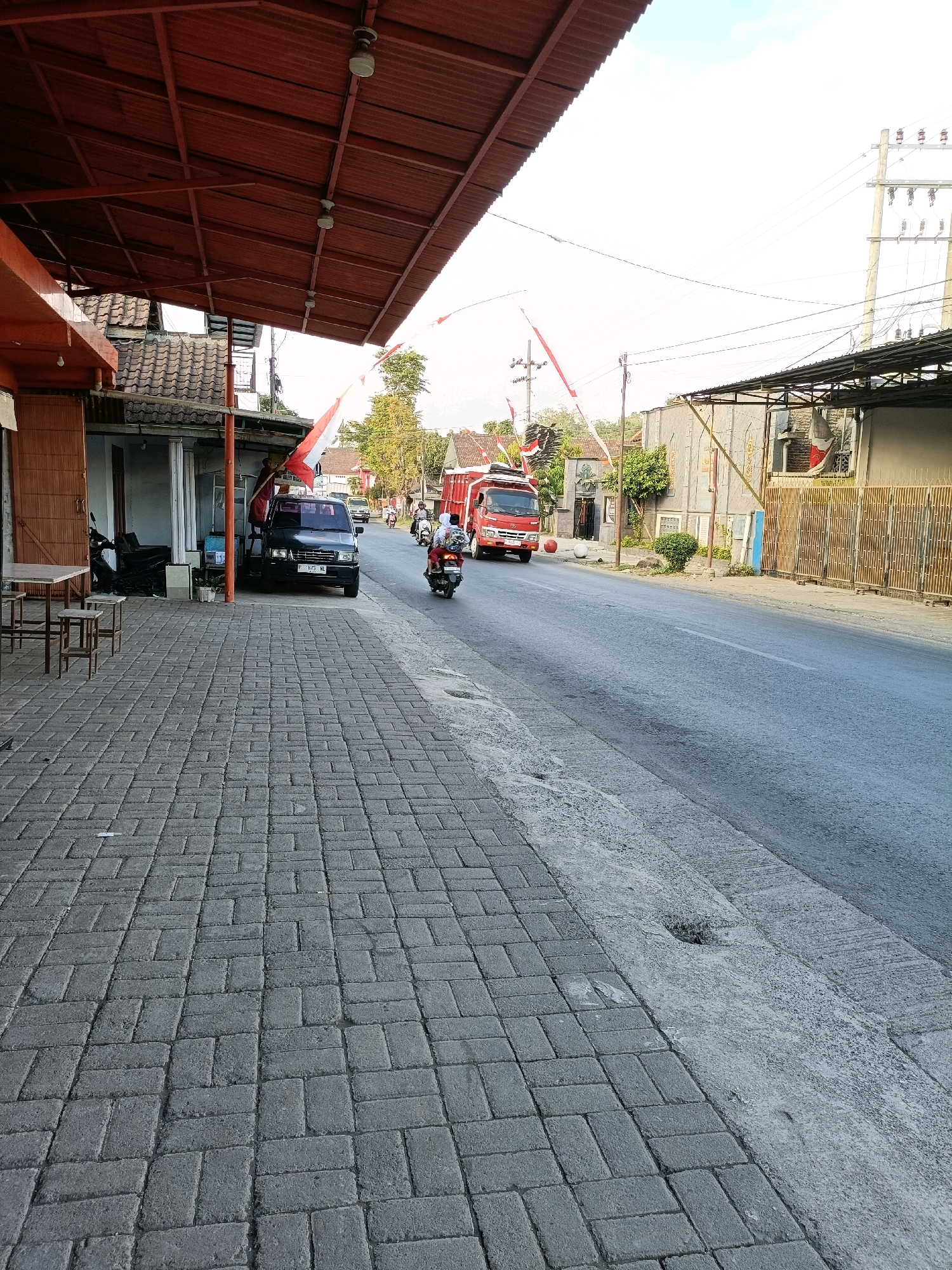 The image depicts a street scene in Tamanan, Indonesia, characterized by its paved sidewalk and asphalt road. On the left side of the street, there is a building with an orange roof overhang, featuring a doorway and a display of tables and stools. A black car is parked in front of the building. A red truck is driving on the road, followed by two motorcycles, one carrying two passengers and the other carrying one. The environment is somewhat rural, with trees and houses visible in the background. The sky is bright and appears to be daytime. The scene is decorated with numerous Indonesian flags, suggesting a national celebration or event.