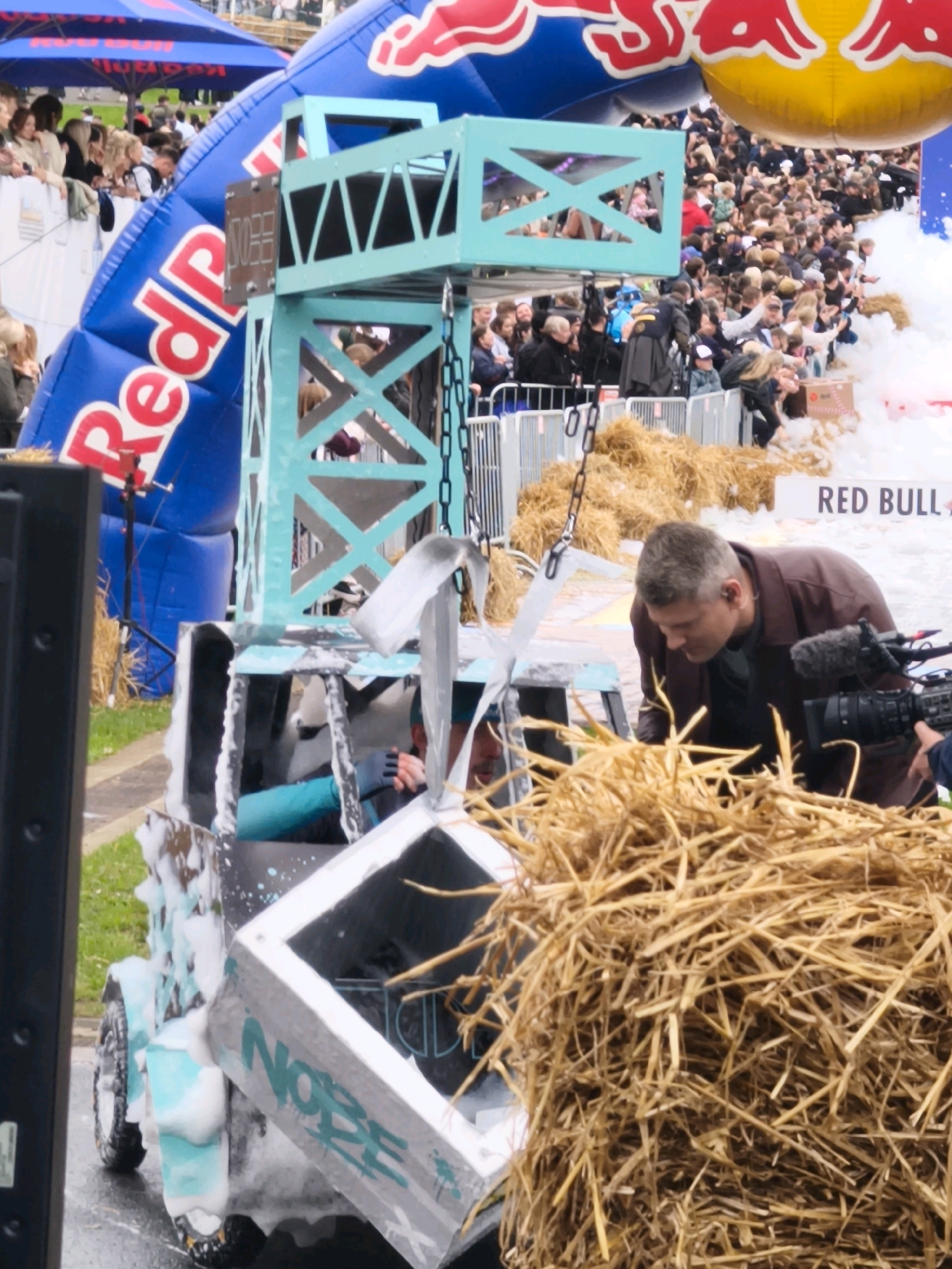 The image depicts an outdoor event, likely a competition or festival, characterized by a large inflatable structure with the "Red Bull" logo prominently displayed. A teal-colored crane-like structure stands near the center, with a vehicle or cart underneath it, partially covered in foam and featuring the word "NOBE" spray-painted on its side. Several individuals are visible, including a man in a brown jacket looking down, seemingly engaged with the vehicle. A crowd of spectators can be seen in the background. The environment appears to be a paved area with grass bordering it, and a significant amount of straw is present in the foreground and around the vehicle. The presence of Red Bull branding suggests a connection to extreme sports or event marketing. There is no indication of the specific location within the city of Tallinn, Estonia.