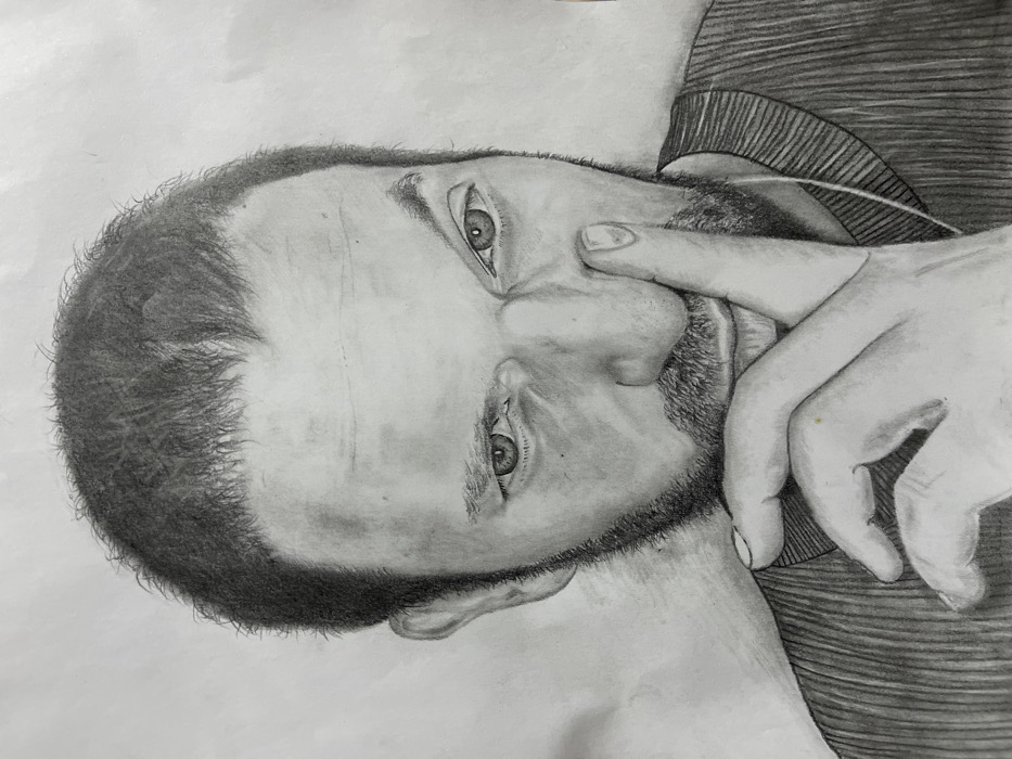 The image is a grayscale pencil portrait drawing depicting an adult male. The subject has short, dark, textured hair and a visible beard or stubble around the jawline and chin. His eyes are open and directed forward. The right hand is raised, with the index finger touching the right side of the nose. The thumb and remaining fingers are partially visible, curled below the chin. The subject is wearing a dark, ribbed garment, likely a sweater or long-sleeved shirt. A thin, light-colored line, consistent with an earbud wire, is visible descending from the right ear area, across the neckline. The background is a plain, light-toned surface without discernible features. The scene is situated in Jos, Nigeria.