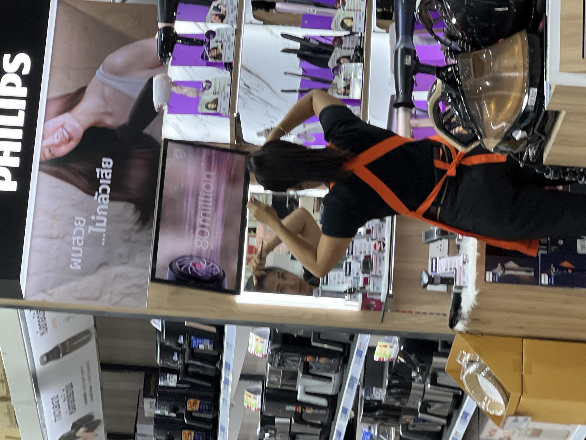 In Baan Chaweng Noi, Thailand, an interior shot captures a retail display in what appears to be an electronics or appliance store. The scene is dominated by shelves stocked with various home appliances, primarily air fryers and hair styling tools. Prominently displayed are Philips brand products, advertised with large text and images. One large screen shows a model with long, dark hair, accompanied by text that reads "ผมสวย...ไม่กลัวเสีย" (Beautiful hair... not afraid of damage). A woman wearing an orange apron over a black t-shirt and black pants is visible from the back, interacting with her hair. She is positioned in front of a mirror, revealing her reflection as she appears to be styling her hair. The shelves showcase multiple hair dryers and what seem to be steam irons, with packaging and product displays arranged in an organized manner. The lighting is bright, typical of a retail environment, illuminating the products and the overall display.