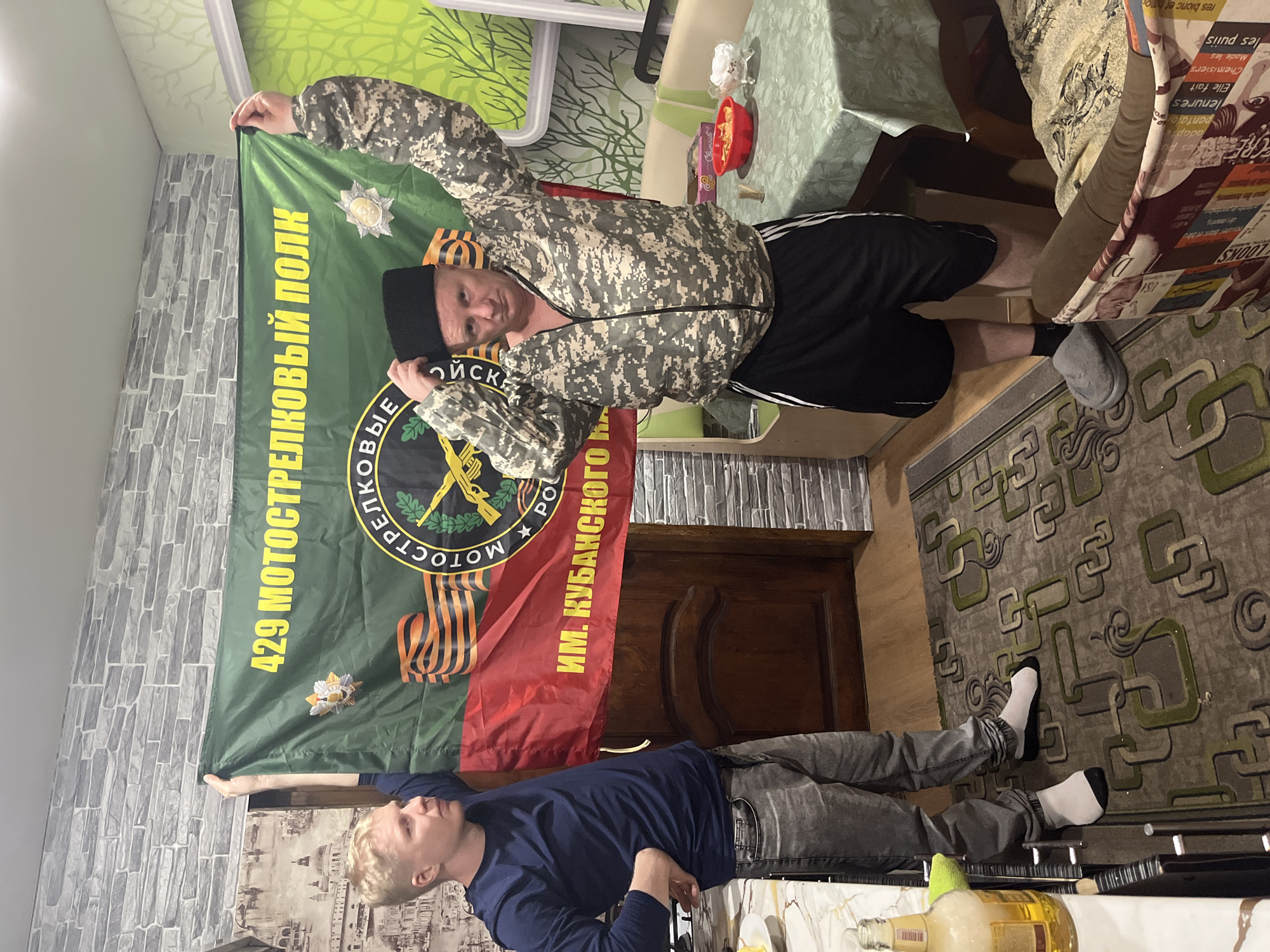 Two men are posing with a flag in what appears to be an indoor setting. One man is holding the top of the flag, wearing a camouflage jacket, black shorts, and a black beanie. The other man is lying on the floor, holding the bottom of the flag, wearing a blue shirt and gray pants. The flag itself is divided into green and red sections, with a circular emblem in the center featuring a rifle and swords. The green section displays yellow text "429 MOTOCTPEIKOBBIЙ ПOJK" (429 Motorized Rifle Regiment). The red section displays yellow text "WM. KYBAHCKOΓO". There is a doorway and a carpet in the background.
Based on the flag displayed and the text on the flag, there are indications that this image could be connected to the 429th Motorized Rifle Regiment, named after the Kuban Cossacks, which may provide information about the location. However, the image alone does not confirm the specific city of Vesyoloye, Russia.