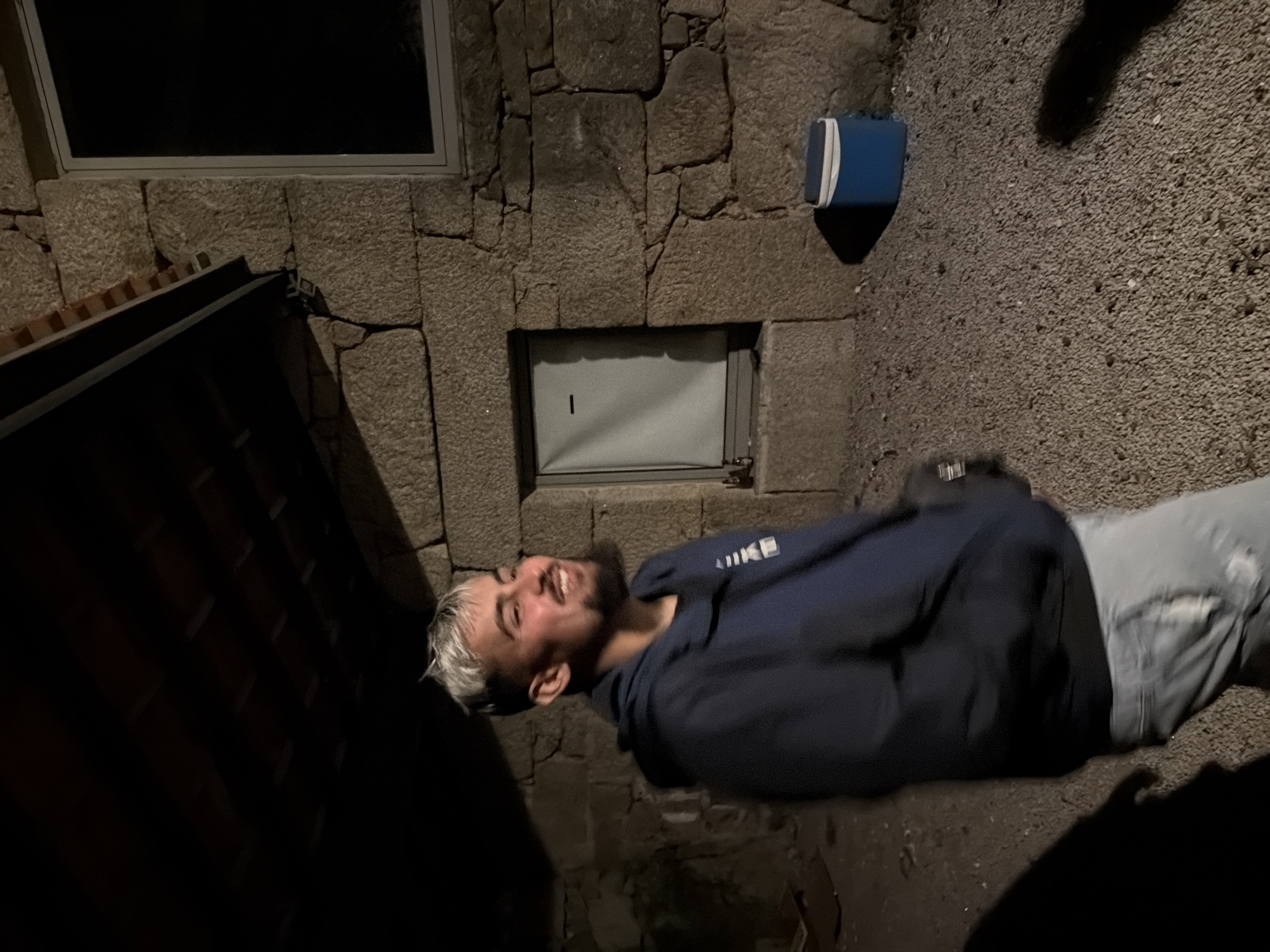 A man is lying on the ground in Vila Real, Portugal, outside a stone building with two windows. The man is wearing a dark blue hoodie and light-colored jeans, and he appears to be smiling. A blue object is located on the stone wall. The area is dimly lit.