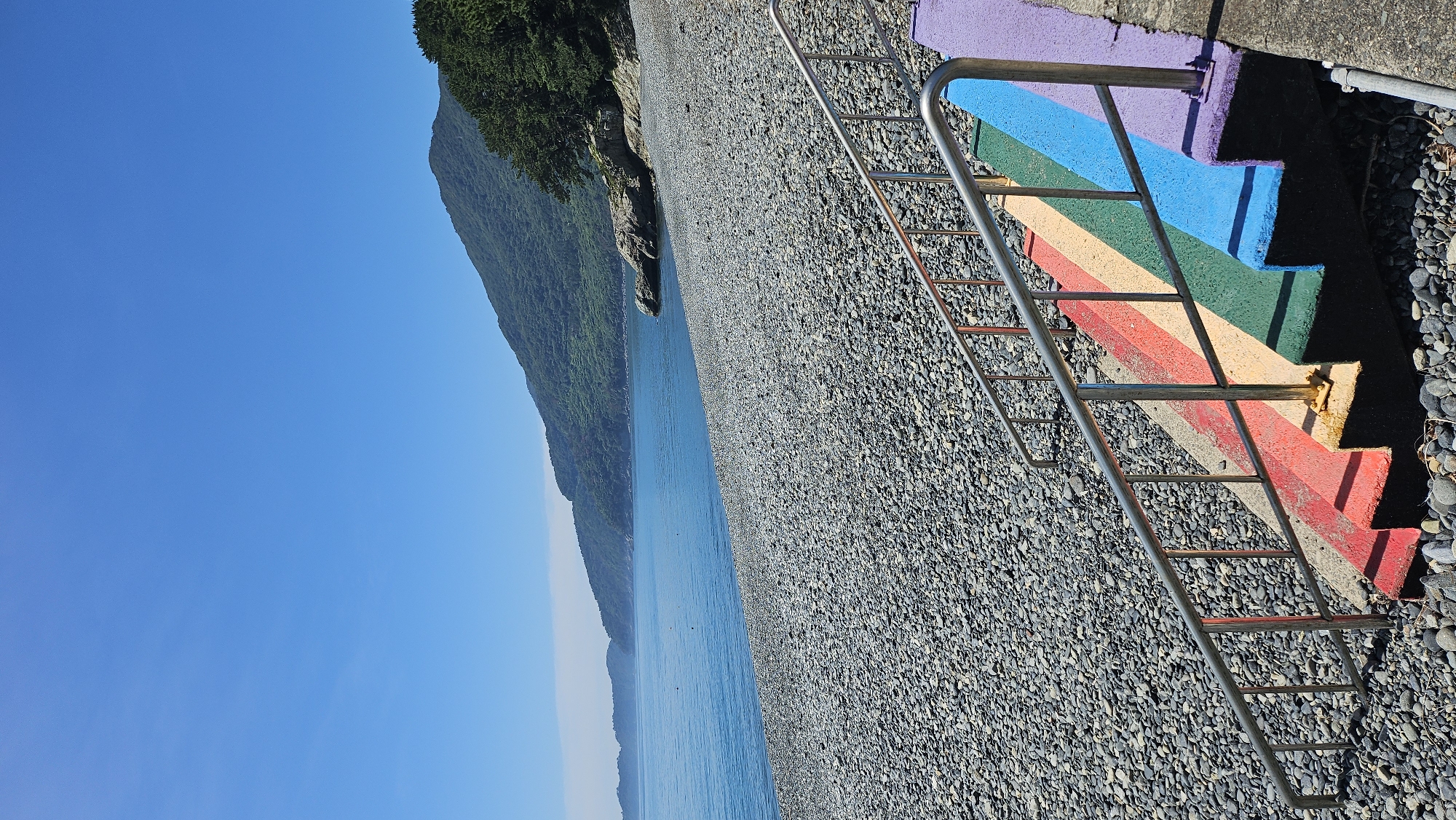A photograph captures a scenic view of a pebble beach with calm, blue water meeting a verdant, mountainous shoreline under a clear, bright blue sky. In the foreground, a set of concrete stairs leading down to the beach is painted with rainbow colors: red, orange, yellow, green, blue, and purple. A silver metal handrail runs along the side of the stairs. The beach is covered in smooth, gray pebbles. The location is identified as a coastal area in Geoje-si, South Korea, likely a public access point to the beach. There are no people visible in the image.