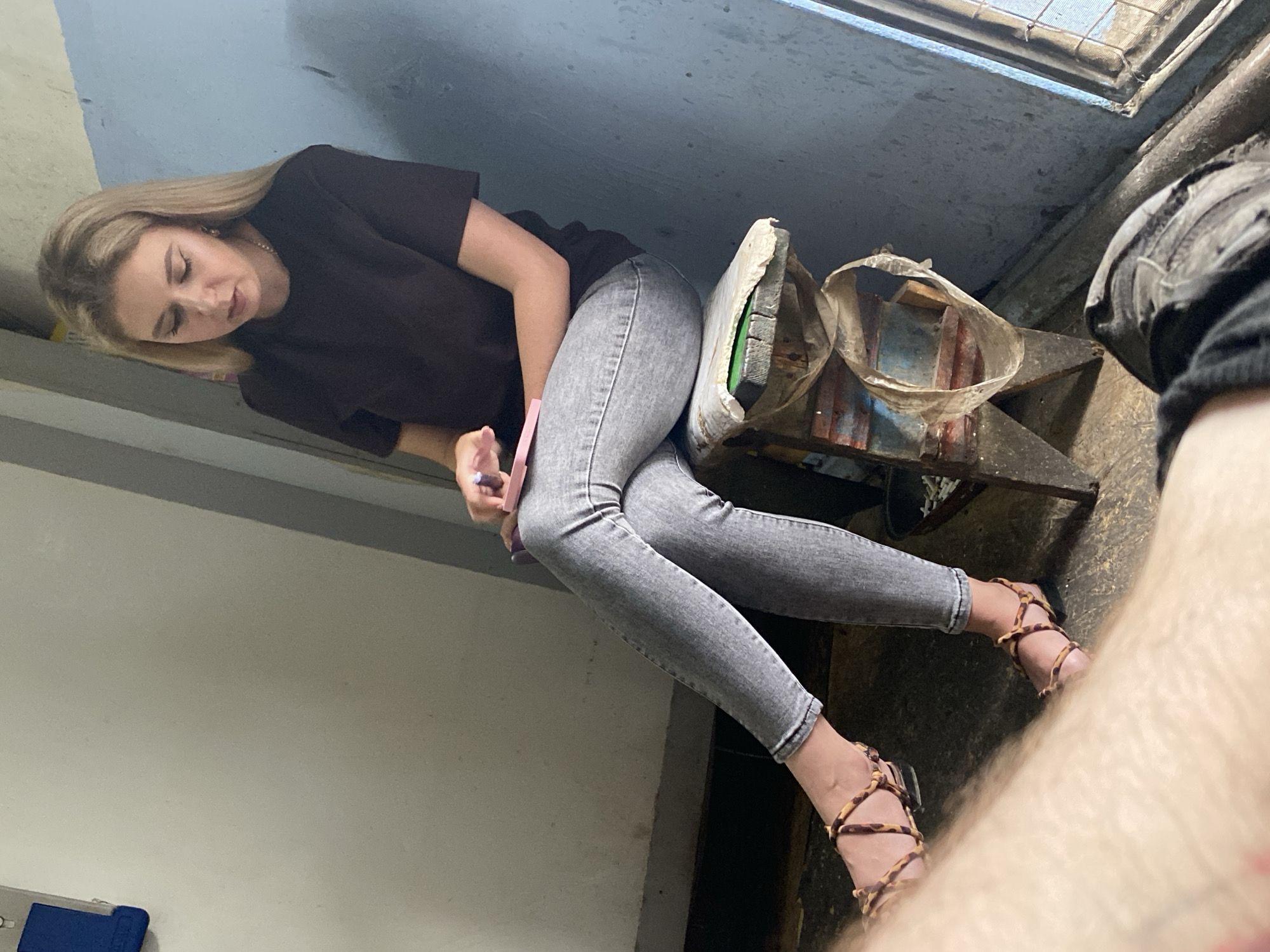 A young woman with blonde hair, wearing a dark t-shirt and grey jeans, sits on a rustic wooden stool. She is holding a pink object, possibly a phone or book, in her hands. Her legs are crossed, and she wears strappy sandals. The background consists of a light-colored wall and a blue painted wall with a window. The scene appears to be indoors in a sparsely furnished area.