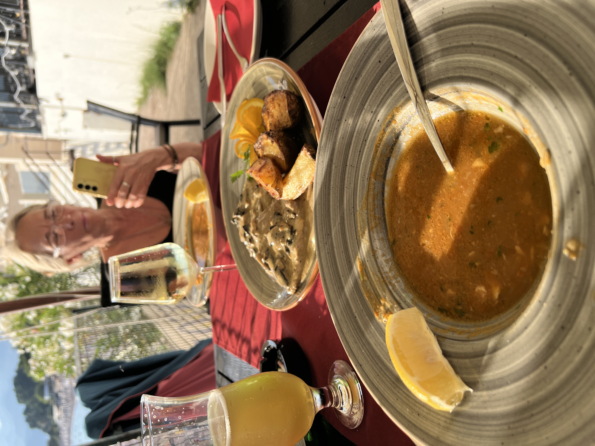 A woman with blonde hair and glasses is seated at a table on an outdoor terrace, holding a yellow smartphone in her right hand and looking at it. She is wearing a black top and has a bracelet on her left wrist. In front of her, on the table, are two plates of food, one with a creamy mushroom dish and roasted potatoes, and another with a partially eaten orange soup with a slice of lemon. There are also two glasses, one with white wine and the other with a yellow beverage, likely beer. The table is covered with a red placemat, and the background shows a glimpse of the sea and buildings in Sveti Stefan, Montenegro. The overall scene depicts a meal at an outdoor restaurant.