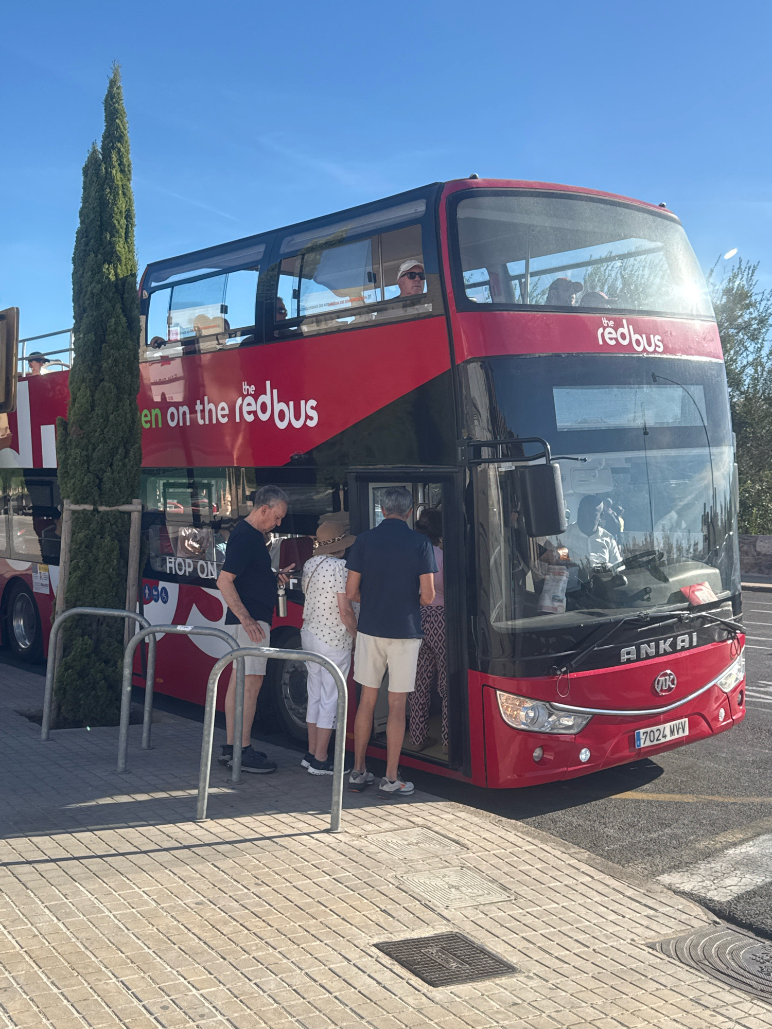 A bright red double-decker bus, identified as "the redbus," is parked on a sidewalk paved with light gray square tiles. The bus has its lower deck door open, and several people are standing near the entrance. One man, wearing a dark blue t-shirt and light-colored shorts, is looking at his phone. Next to him, a woman in a patterned dress and a straw hat is partially visible. Another man in a dark blue t-shirt and light shorts stands behind her.
The bus has "Hop On Hop Off" signage visible on its side, and the word "ANKAI" is emblazoned on the front. The license plate reads "7024 MVV". A tall, slender, dark green tree stands to the left of the bus. The background reveals a clear blue sky and some greenery. The overall scene suggests a sunny day, possibly a tourist stop in the city of València, Spain, given the presence of a hop-on hop-off bus service.