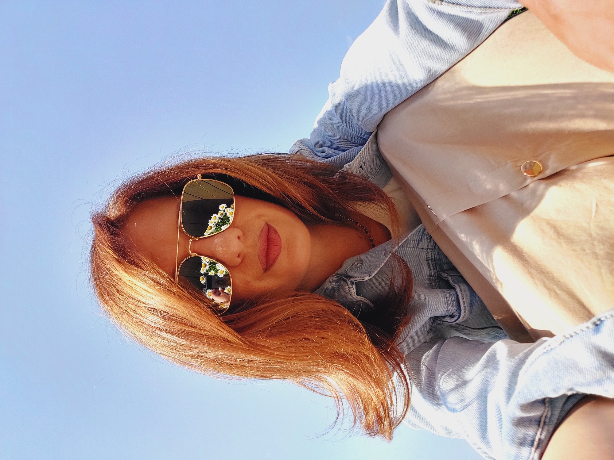A low-angle selfie is taken from a worm's eye view, looking up at a woman with auburn hair and fair skin against a clear blue sky. She is wearing aviator-style sunglasses with gold frames, and the lenses reflect a cluster of white daisies. A denim jacket is layered over a beige top, and her legs are crossed. The scene is shot outdoors with natural light, and shadows are cast across her clothing, suggesting it is daytime. The backdrop is a uniform, cloudless blue sky. There are no discernible landmarks or elements that would pinpoint the location to Sofia, Bulgaria.
