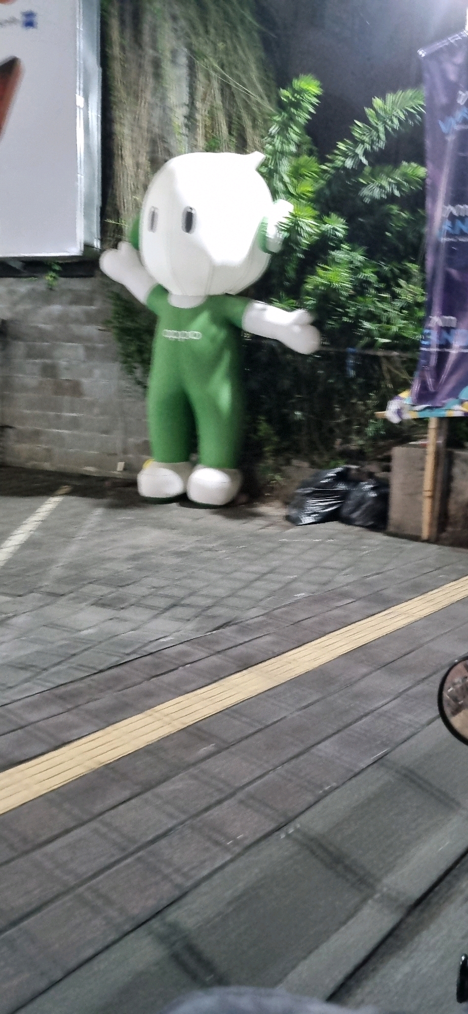 A full-body, inflatable mascot, approximately 7 feet tall, stands on a gray, brick-patterned walkway in Denpasar, Indonesia. The mascot is white and round with black oval eyes and a small, green, pointed protrusion on its head. It wears a full-body, green suit with white accents on the hands and feet. The suit has a white, circular emblem on the chest. The mascot's arms are extended outwards and slightly upwards.
Behind the mascot, a brick wall is visible, partially covered by green foliage. To the right of the mascot, a dark blue banner hangs from a wooden pole, with illegible white text. Two black garbage bags are placed on the ground to the right of the mascot.
The walkway is paved with large, gray tiles arranged in a pattern. A raised, yellow tactile paving strip runs diagonally across the walkway in the foreground. The scene appears to be outdoors at night, with artificial lighting illuminating the mascot and its surroundings. In the lower right corner, a portion of a motorcycle's rearview mirror is visible, reflecting the scene. The overall environment suggests an urban setting, likely a public area or event space in Denpasar.