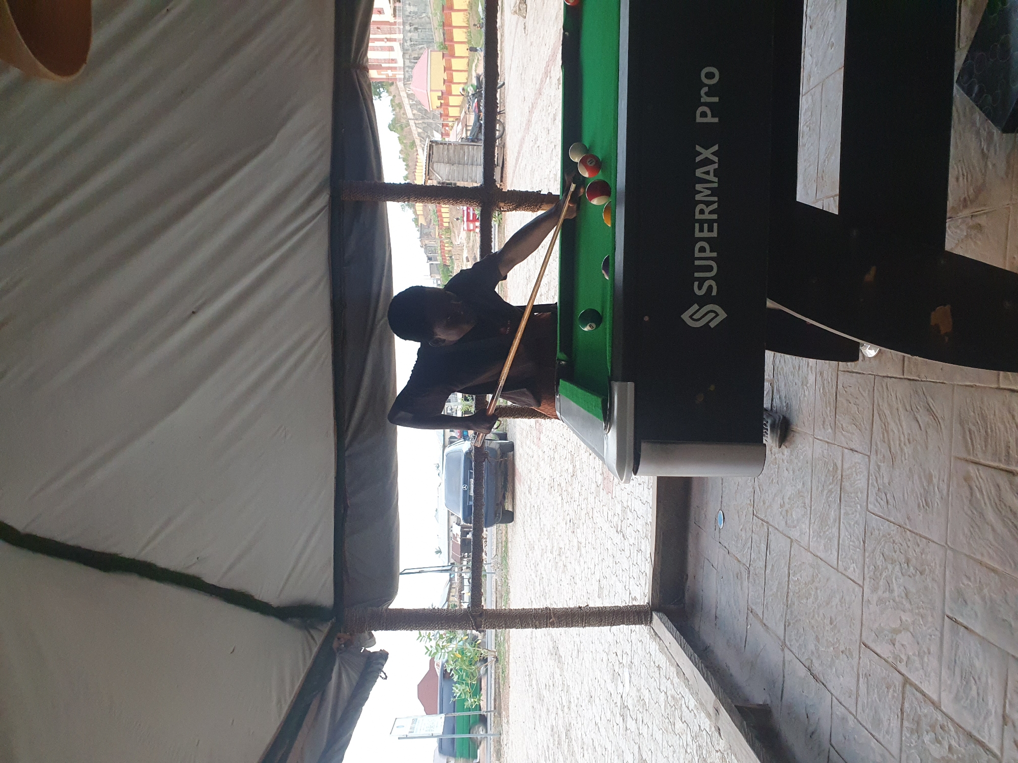 A man is playing pool on a "SUPERMAX Pro" pool table. He is indoors, in an open-walled structure with a white tent roof. Outside, across a partially obscured street, are buildings with a red roof and parked cars. The location is Unknown, Unknown.