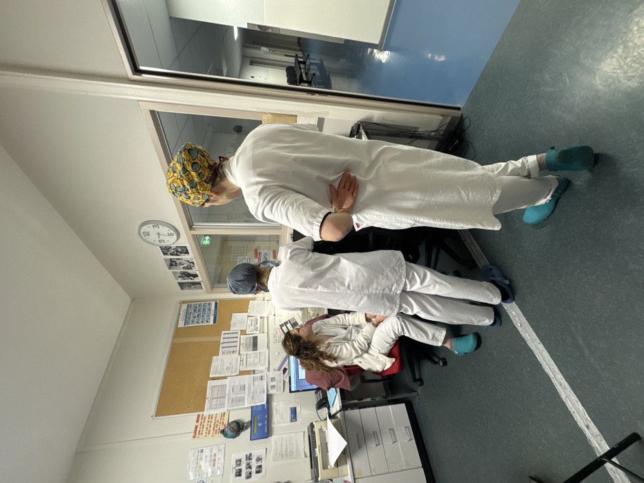 Three people in medical attire, likely doctors or nurses, are in a room. Two stand near a window, looking out. One wears a patterned surgical cap, the other a solid blue one. The third individual sits at a desk with a computer. Various papers and notices are pinned to a corkboard behind the desk. A clock is visible on the wall. The room appears to be in a hospital or medical facility. All three wear matching light-colored scrubs and blue clogs.