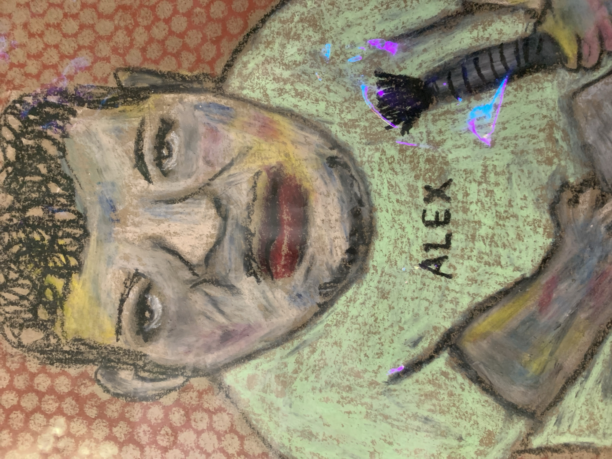 This is a pastel drawing of a man named Alex. He is wearing a light green t-shirt and his name is written in black letters on the shirt. He has dark, curly hair and his face is rendered with a mix of colors, including gray, yellow, and red. He appears to be holding a dark object with purple and blue accents in his right hand, and his left hand is partially visible. The background is a reddish-orange color with a repeating pattern of small, light-colored circles. The artwork has a rough, textured appearance, typical of pastel on a textured surface. There is no discernible indication of the city of Kirov, Russia within the image.