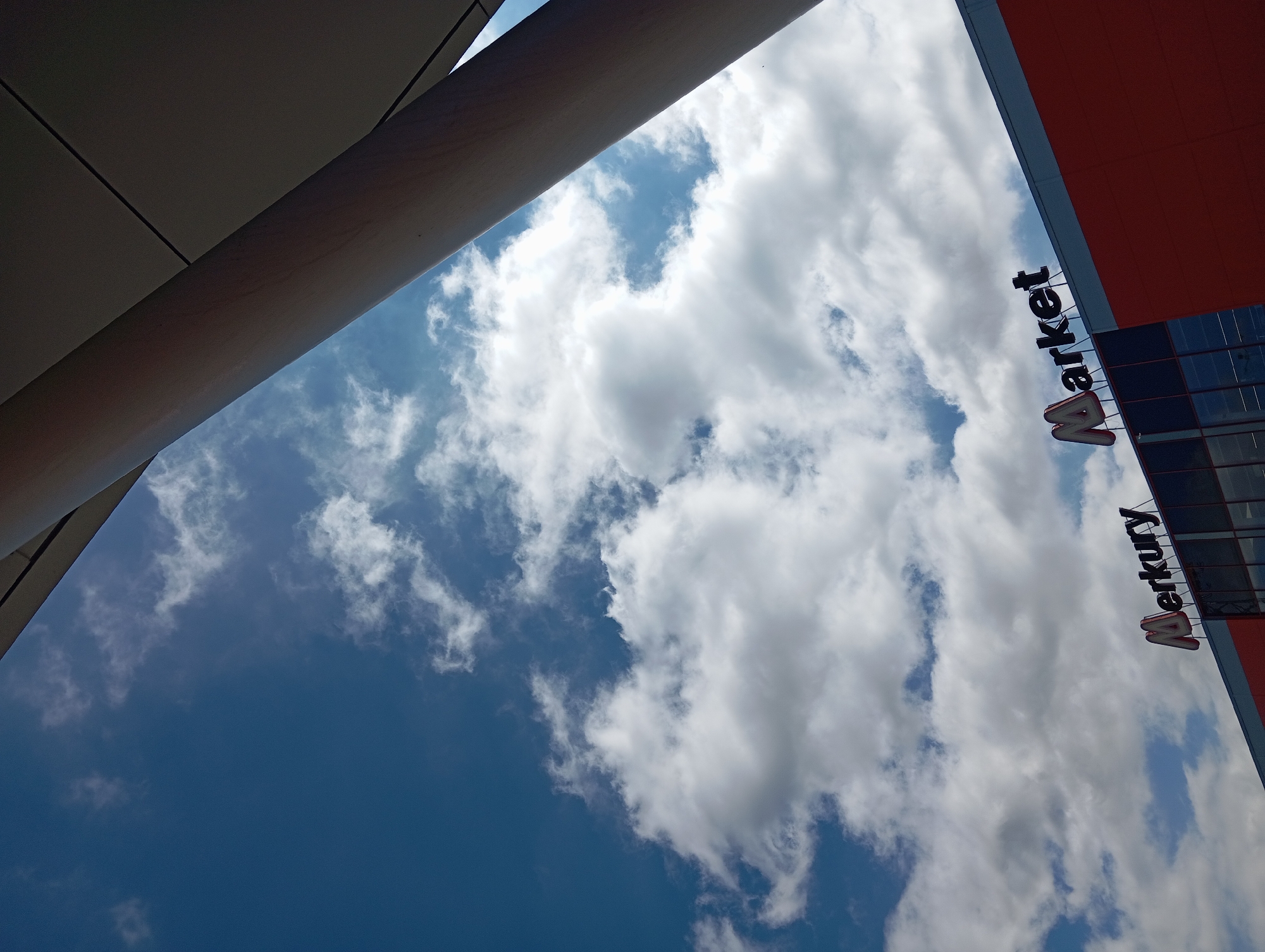 This image captures an upward view of the "Merkury Market" building in Nitra, Slovakia, under a partly cloudy blue sky. The lower portion of the building visible features a red-orange facade and dark reflective windows. Prominently displayed on the building's roofline is the "Merkury Market" sign; "Merkury" is rendered in stylized red and white letters, while "Market" appears in black letters. A large, light-colored cylindrical architectural column or support beam extends diagonally from the upper right toward the center-right, partially framing the building and sky. Another flat, dark grey structural element is visible in the extreme upper right corner. The sky is bright blue with numerous white and light grey cumulus clouds scattered throughout. No people, vehicles, or specific actions are present in the frame.