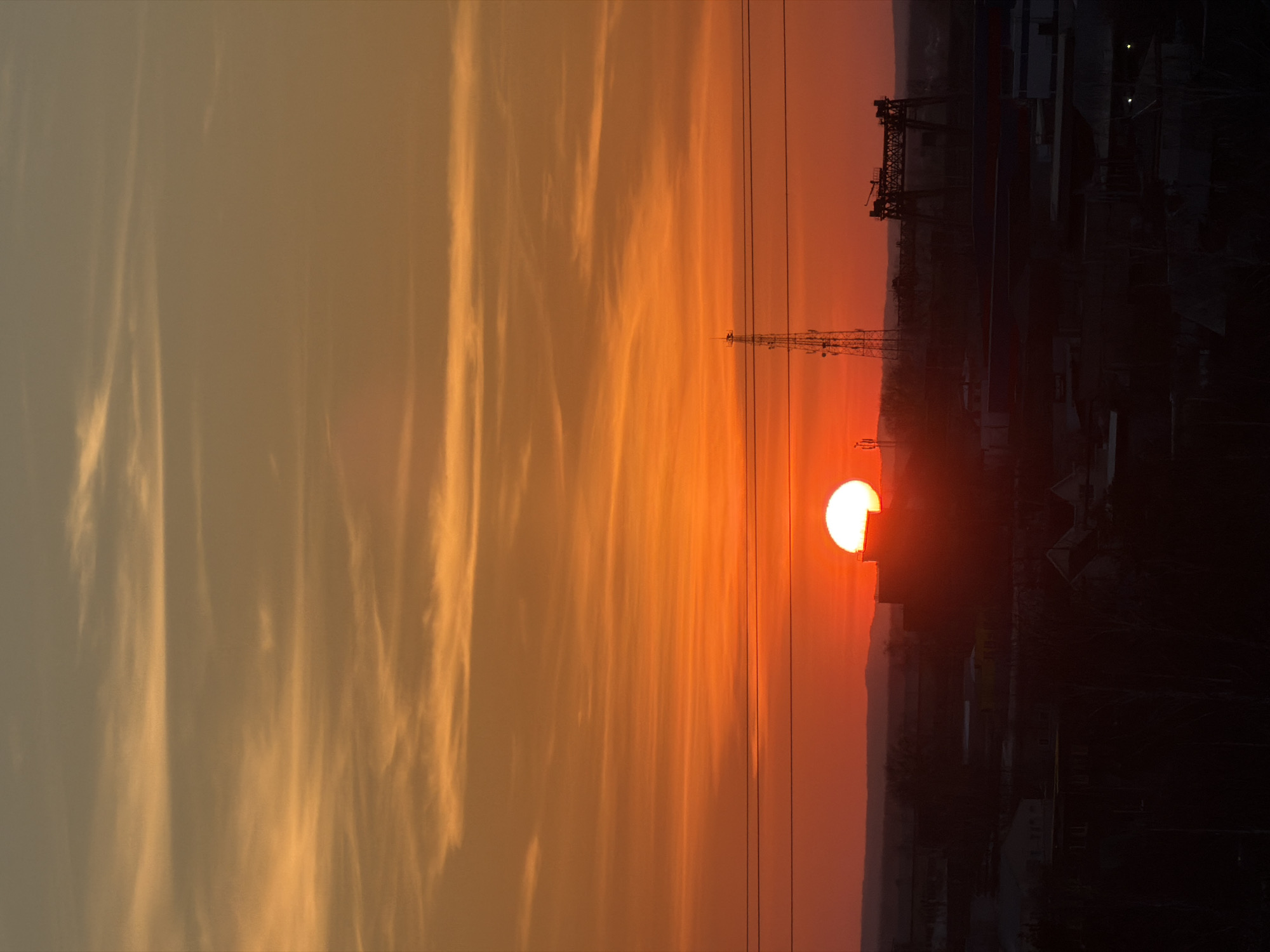 The image depicts a sunset in Chaikovsky, Russia. The sun is a bright orange orb, partially obscured by the horizon. Silhouetted against the sunset are industrial structures, including a gantry crane and power lines. The sky is filled with wispy clouds, tinged orange by the setting sun.