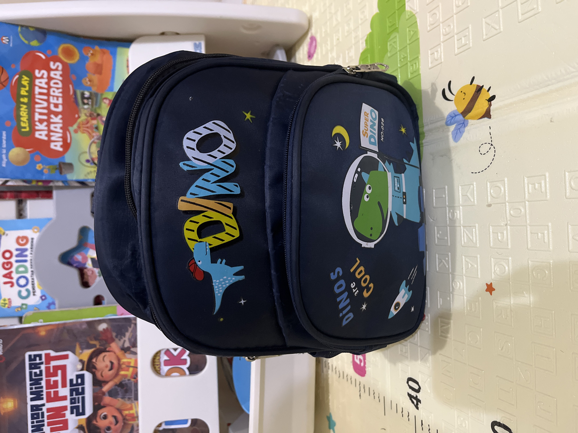 A dark blue backpack with a dinosaur theme is placed on a play mat. The backpack features colorful applique decorations of dinosaurs, rockets, and stars, as well as the text "DINO" and "Dinos are COOL". Below the main design, there is a smaller compartment with a "SUPER DINO" label and the number "NO:028". In the background, several children's books are visible, including one titled "JAGO CODING" and another labeled "LEARN & PLAY AKTIVITAS ANAK CERDAS". A small bee sticker is also visible on the play mat to the right of the backpack. The play mat itself has raised letters and numbers, and a ruler marking along the left edge. The scene appears to be an indoor setting, possibly a child's room or play area. There are no people visible in the image. The lighting suggests it is daytime. The overall impression is one of a child-oriented environment. The image does not contain any information that would suggest a specific location in the city of East Jakarta, Indonesia.