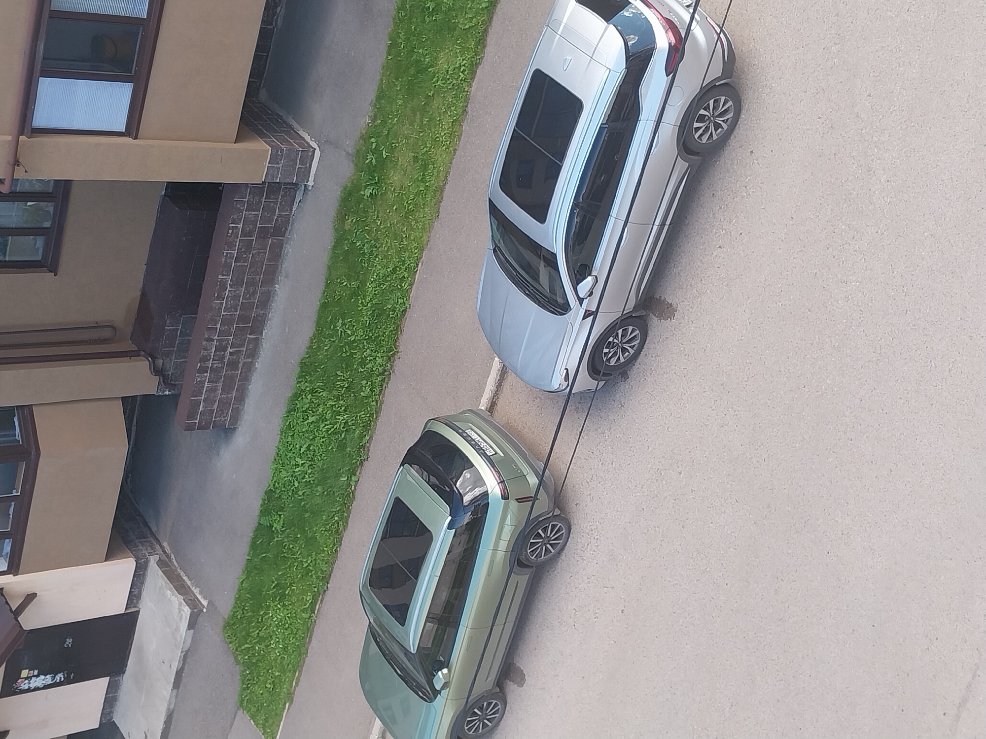 In the city of Salavat, Russia, two SUVs are parked on an asphalt road adjacent to a building with a light brown facade and a brown roof. A thin strip of green grass runs along the side of the road, separating it from the building's foundation, which is decorated with dark brickwork. The first SUV, a light green vehicle, is parked on the left, facing away from the camera. A black towing cable extends from its rear bumper to the front bumper of the second SUV, a silver vehicle, which is parked on the right, also facing away from the camera. Both vehicles have sunroofs.